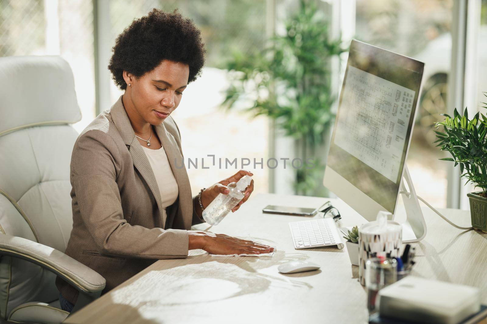 Shot of an attractive African businesswoman sitting alone and using antiseptic gel to disinfect office desk during corona virus pandemic.