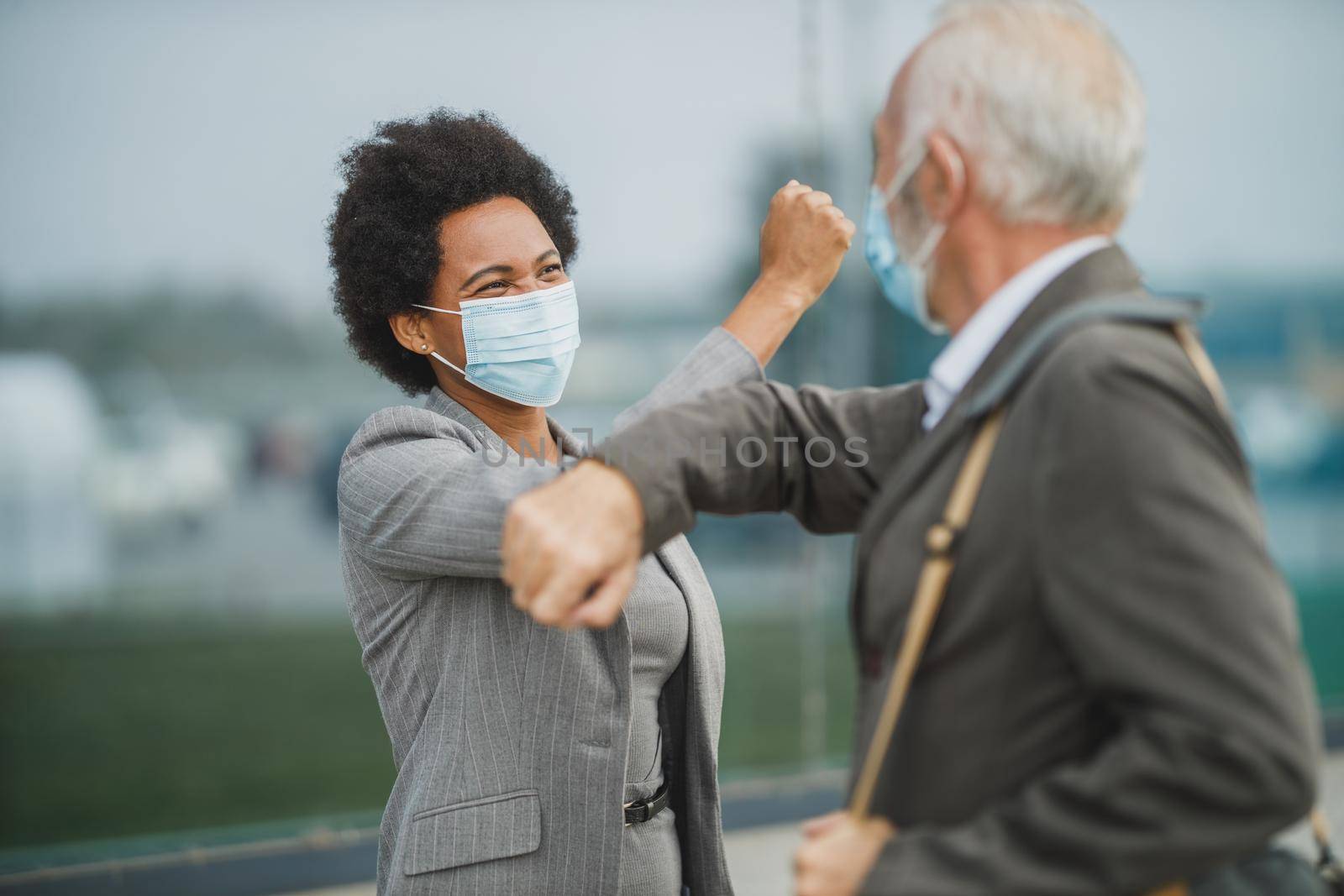 Shot of successful black businesswoman and her senior male colleague using protective mask and greeting with bumping elbows outdoors during COVID-19 pandemic to avoid handshakes.