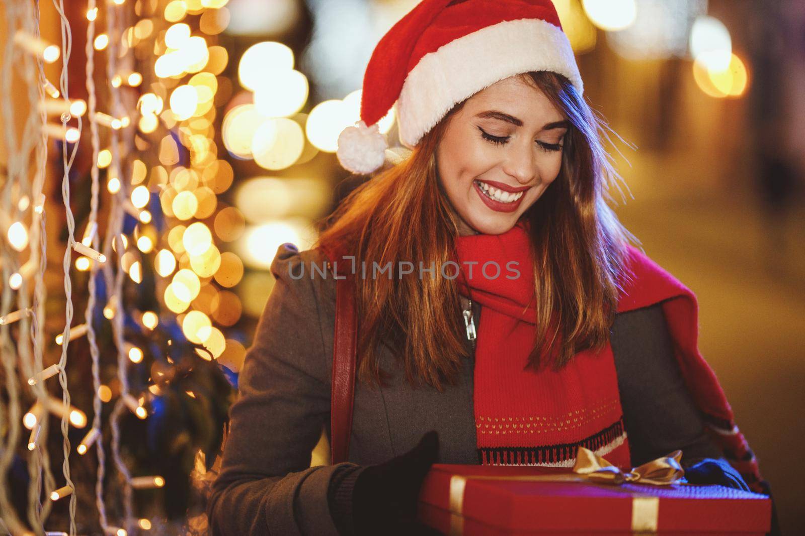 Shoot of a smiling young woman with red present box is having fun in the city street at Christmas time.