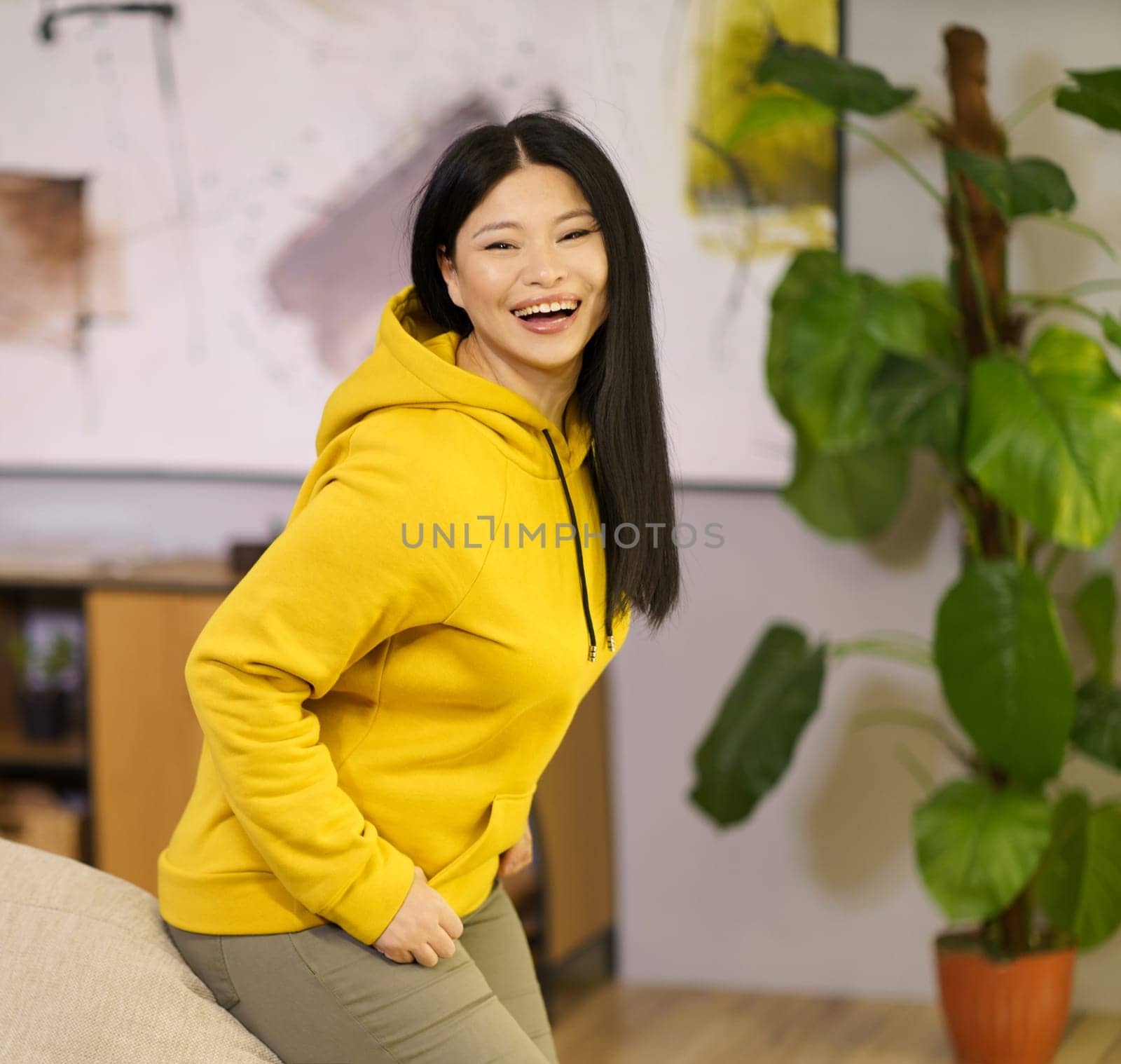 Happy Asian woman is leaning on the sofa and smiling at home. The image represents the concept of a happy and healthy lifestyle with a focus on biohacking by LipikStockMedia