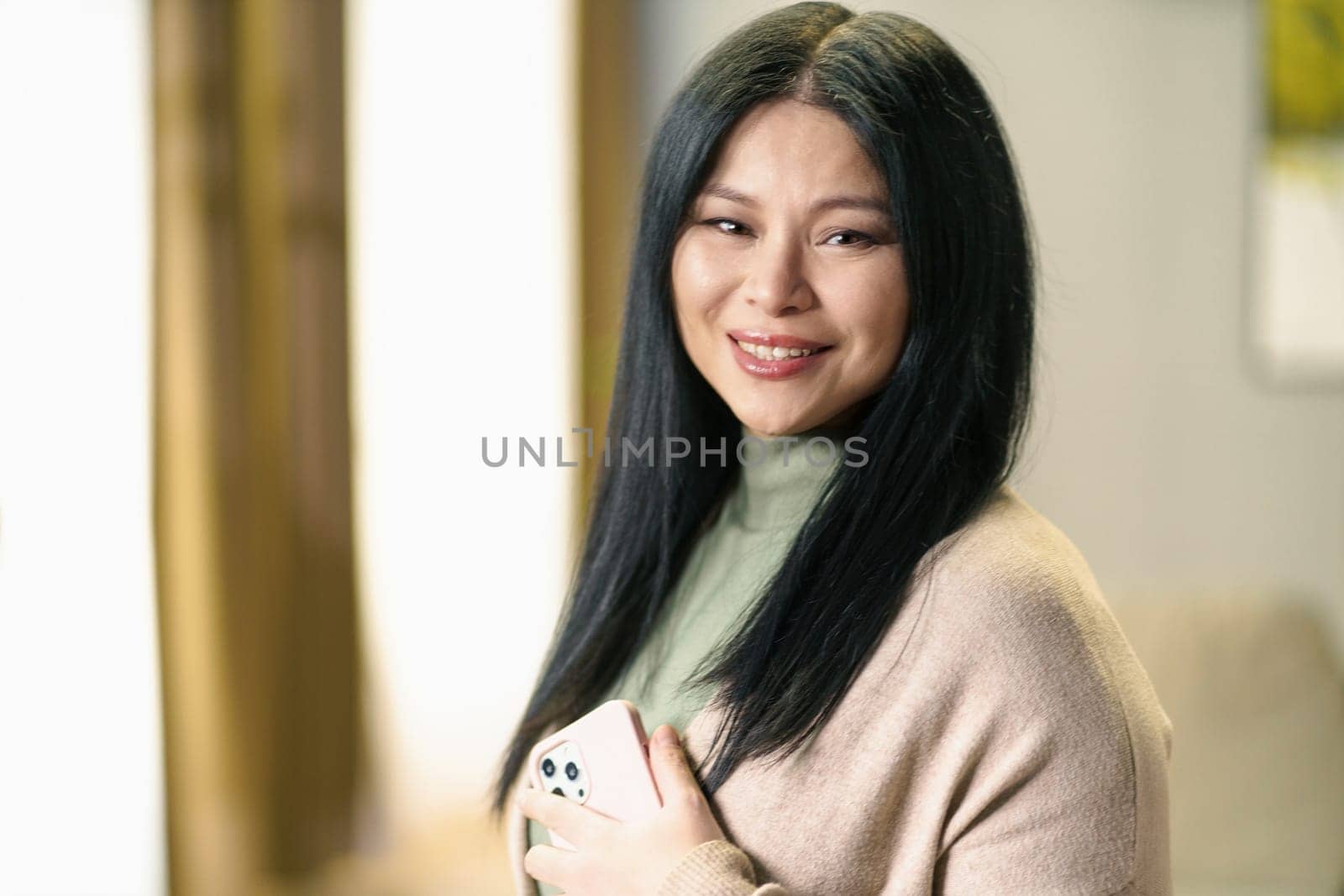 Mature and positive Asian woman with thick, luscious hair holds her smartphone in her hand, smiling with contentment and confidenced by LipikStockMedia