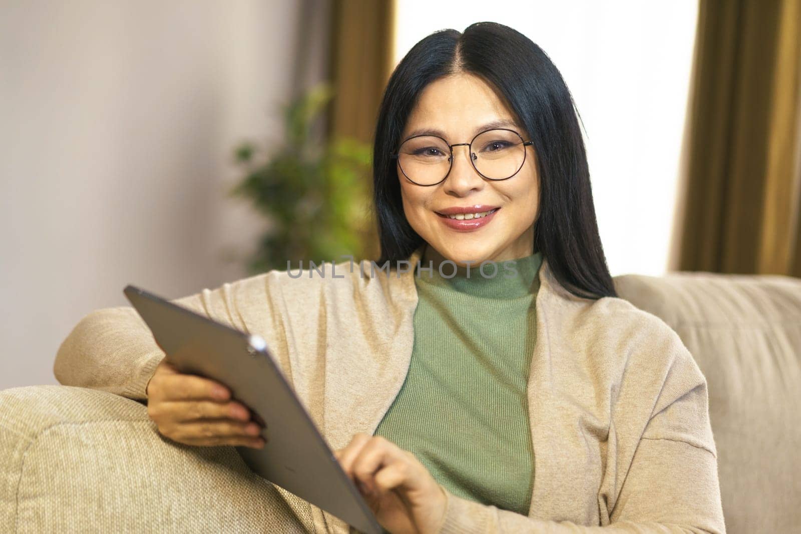 Confident middle-aged Asian woman enjoys moment of relaxation at home, browsing internet on her tablet with ease and comfort by LipikStockMedia
