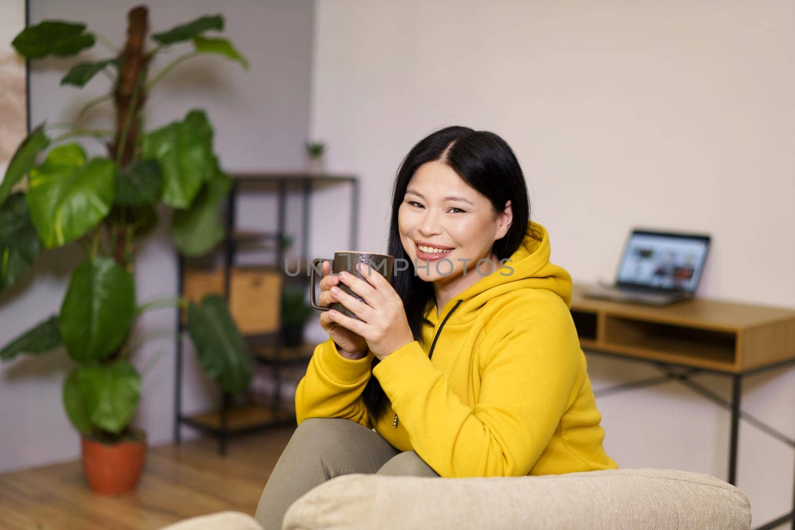 Cute Asian woman takes break from her online work at home to enjoy a soothing cup of tea. The image captures the balance of work and leisure in her comfortable indoor space, surrounded by the calm and serenity of nature. by LipikStockMedia