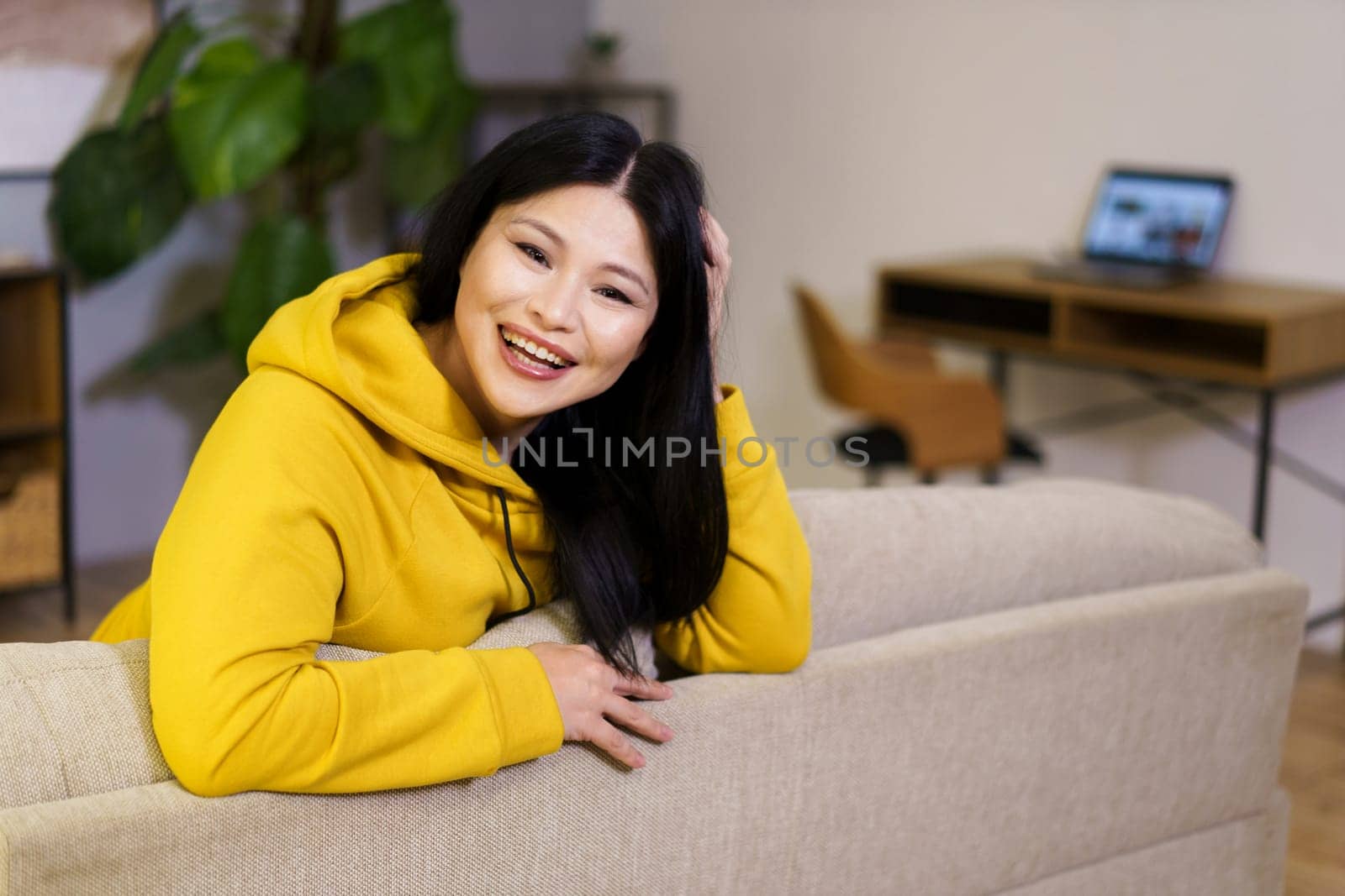 Beautiful middle-aged Asian woman is captured resting on a sofa in her home, surrounded by the comfort and coziness of her indoor space. The background reveals a desktop with a laptop, symbolizing the balance of work and leisure in her lifestyle. High quality photo