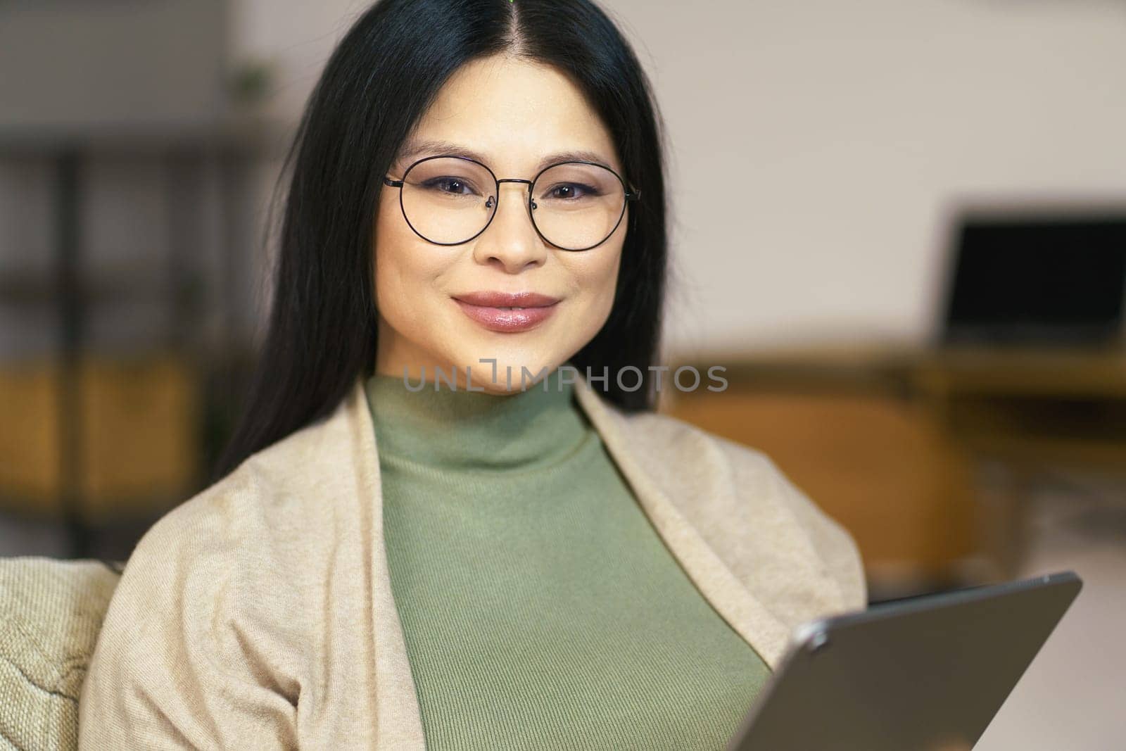 cheerful middle-aged Asian woman wearing glasses enjoys browsing internet on her tablet while sitting comfortably at home by LipikStockMedia