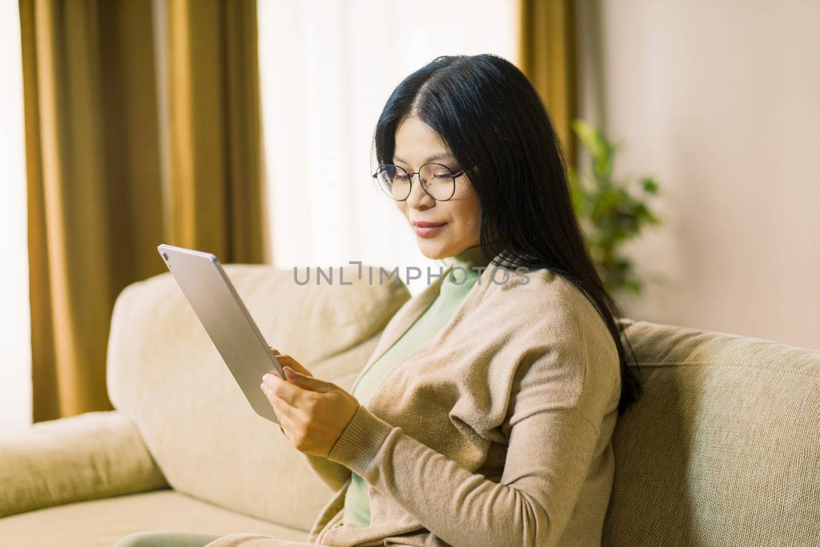 Mature Asian woman wearing glasses is deeply focused on browsing internet on her tablet while comfortably seated in the interior of her home. High quality photo