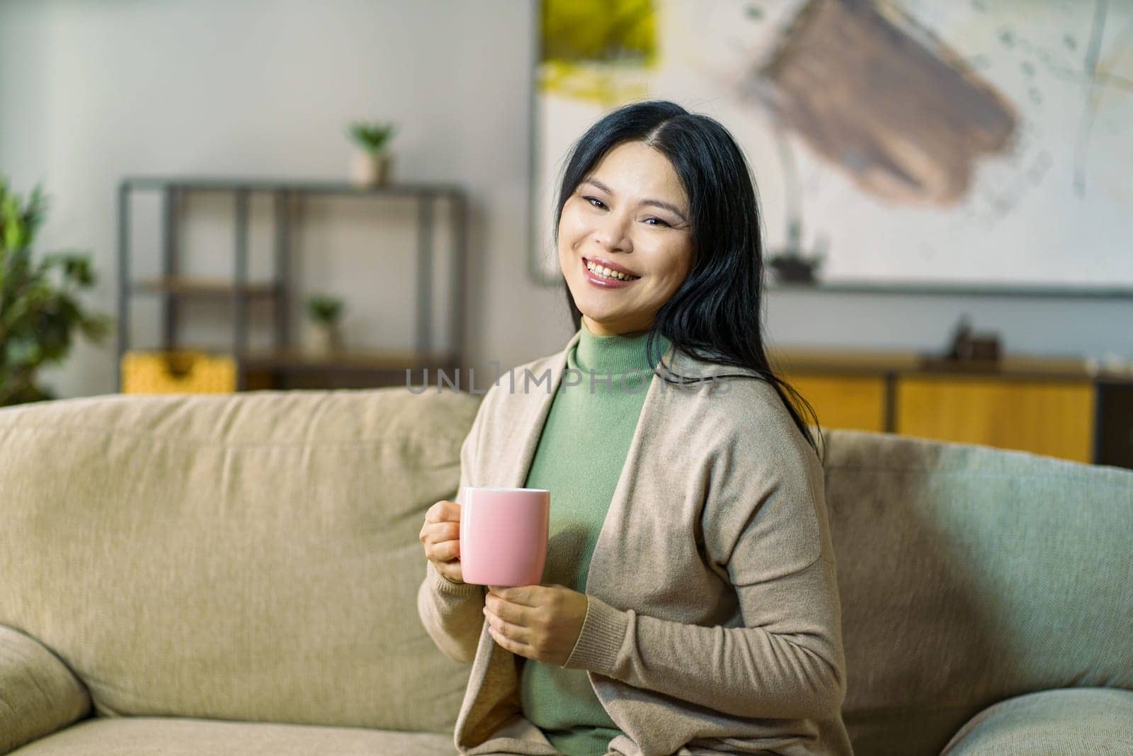 mature Asian woman enjoys a hot cup of tea while relaxing on her cozy living room sofa. High quality photo