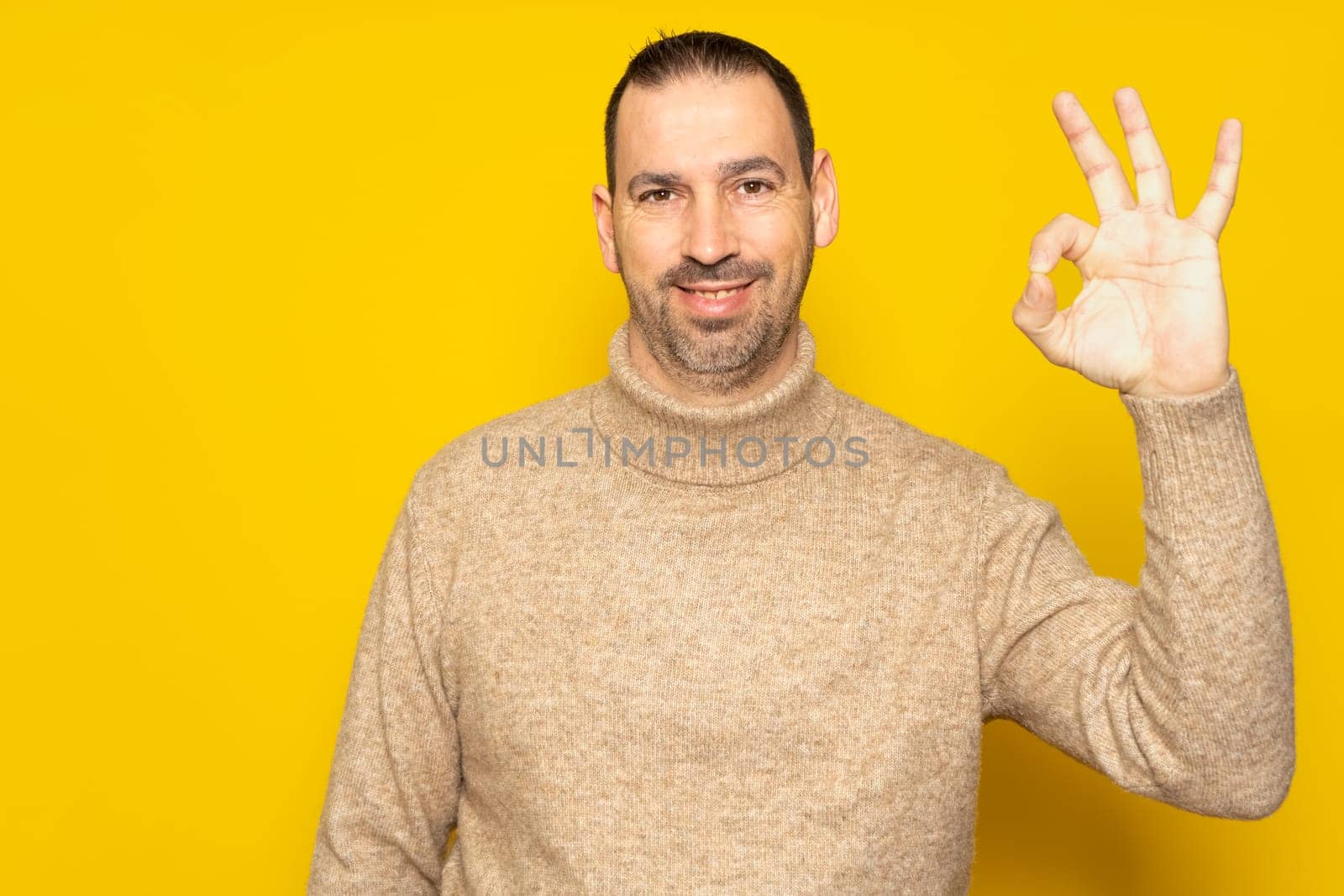 Bearded hispanic man in his 40s wearing a beige turtleneck making the ok gesture while smiling at the camera, isolated over yellow background