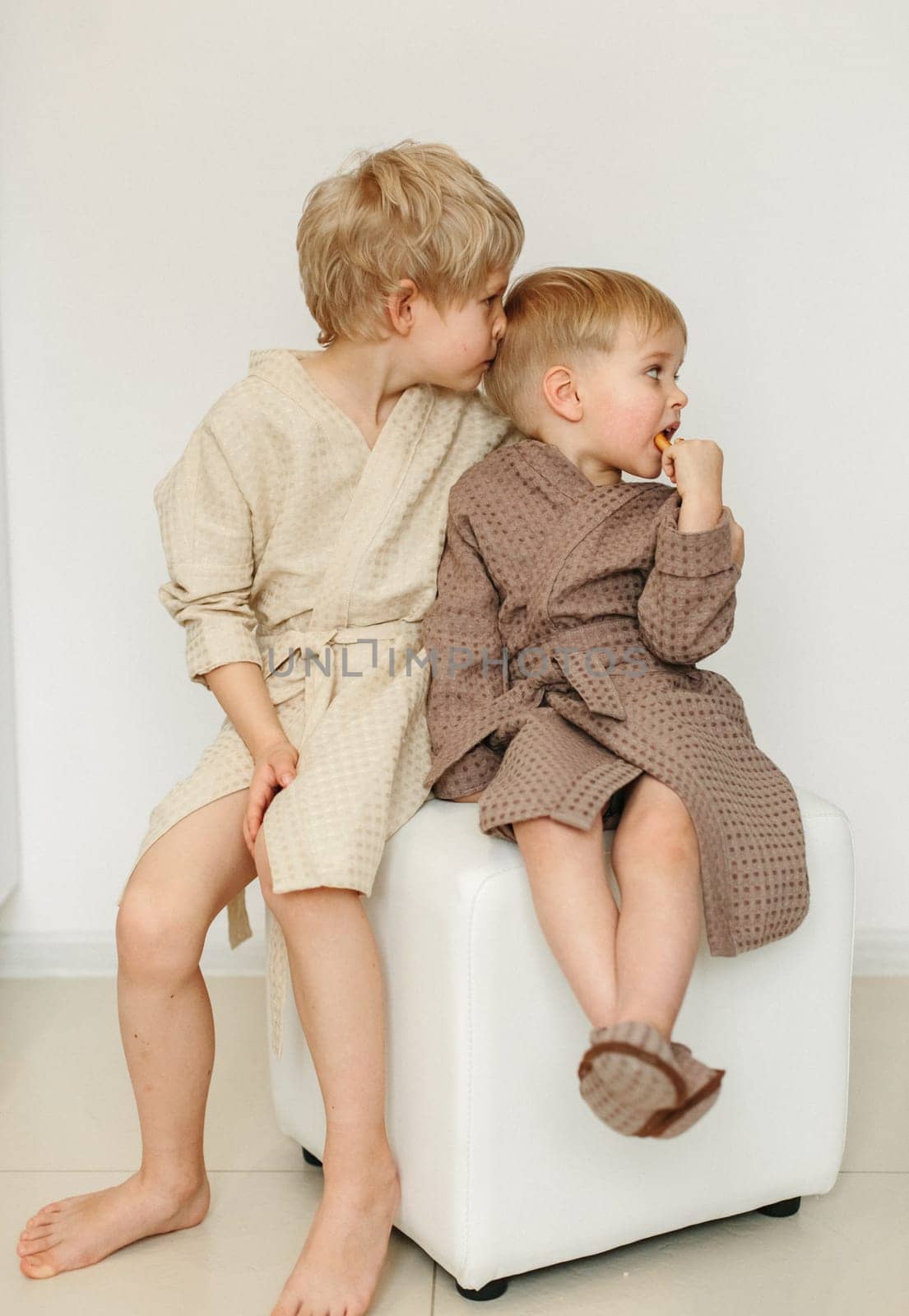 Two boys in bathrobes are sitting on a pouf in a white room. The elder kisses the younger on the back of the head.