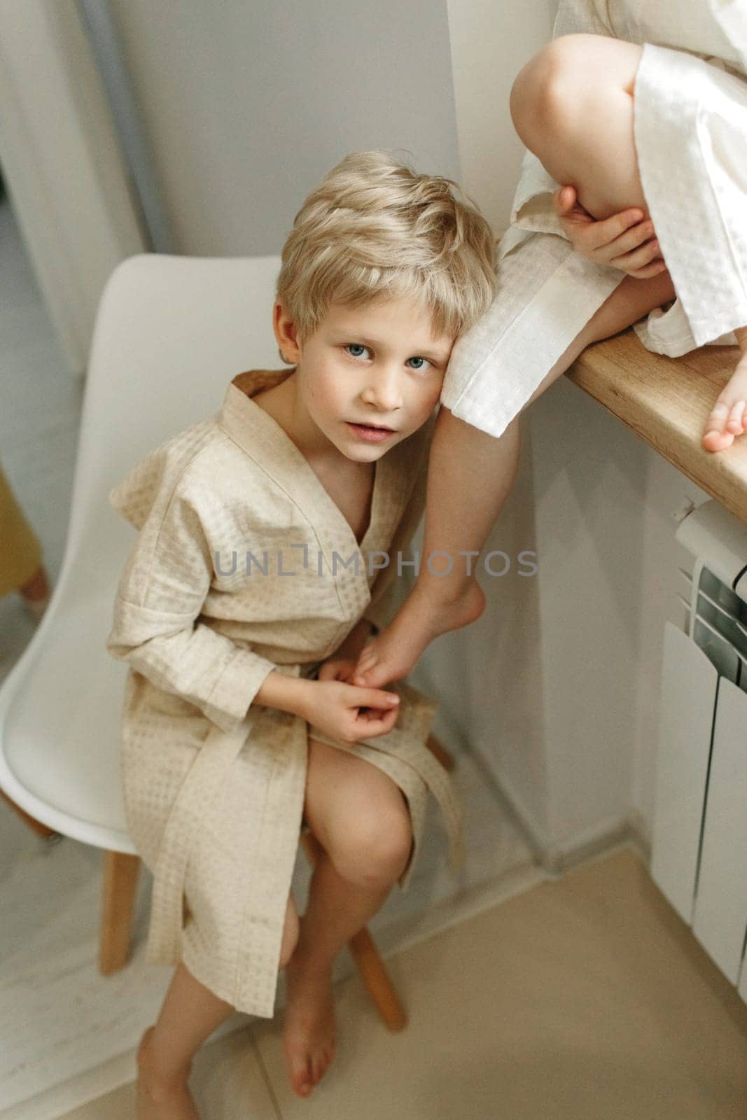 Portrait of a blond boy in a dressing gown, who sits on a chair and leans on his brother's leg.