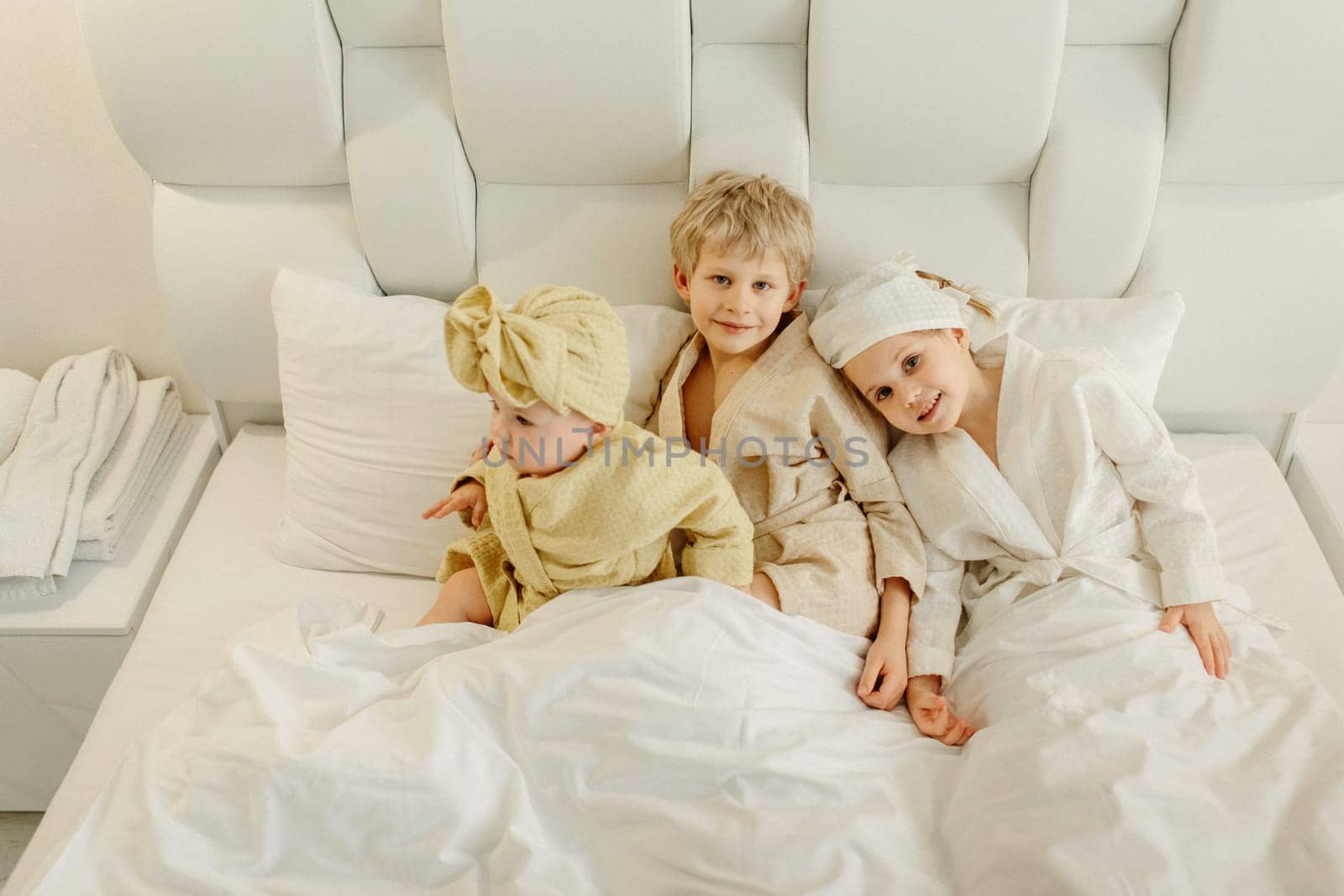 Children lie in a white bed in bathrobes after taking a bath, smiling and looking at the camera. View from above.