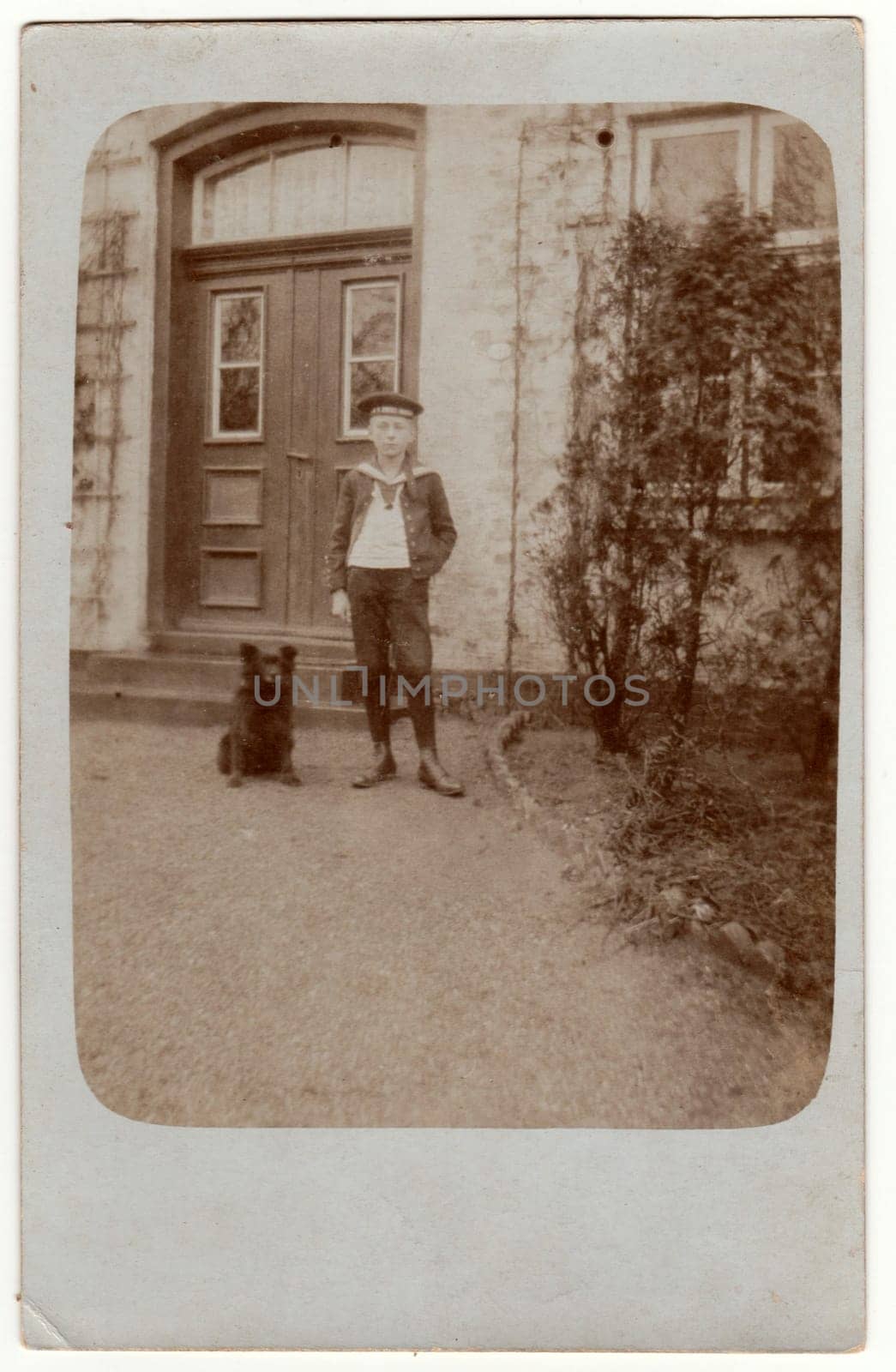 Vintage photo shows boy poses in front of house. Boy wears a sailor costume and stands next to dog. Retro black and white photography. Circa 1920s. by roman_nerud