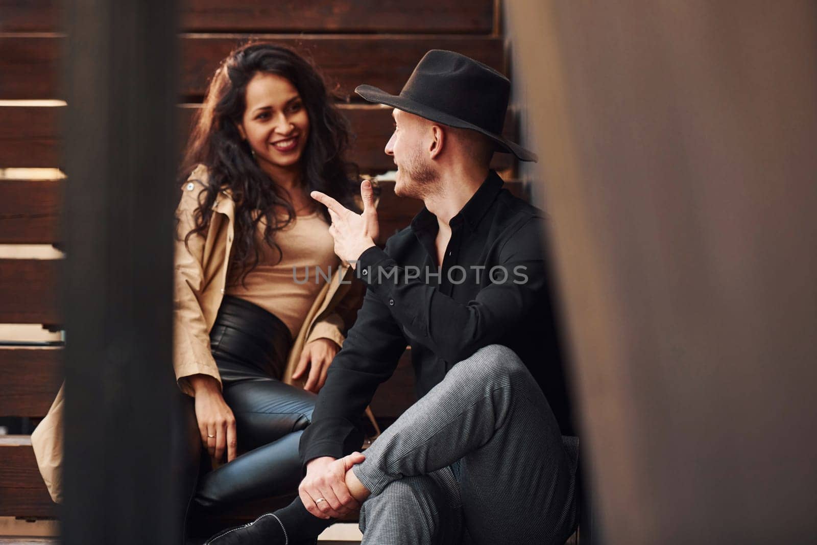 Cheerful brunette in black leggings sitting on wooden stairs with her man in hat.