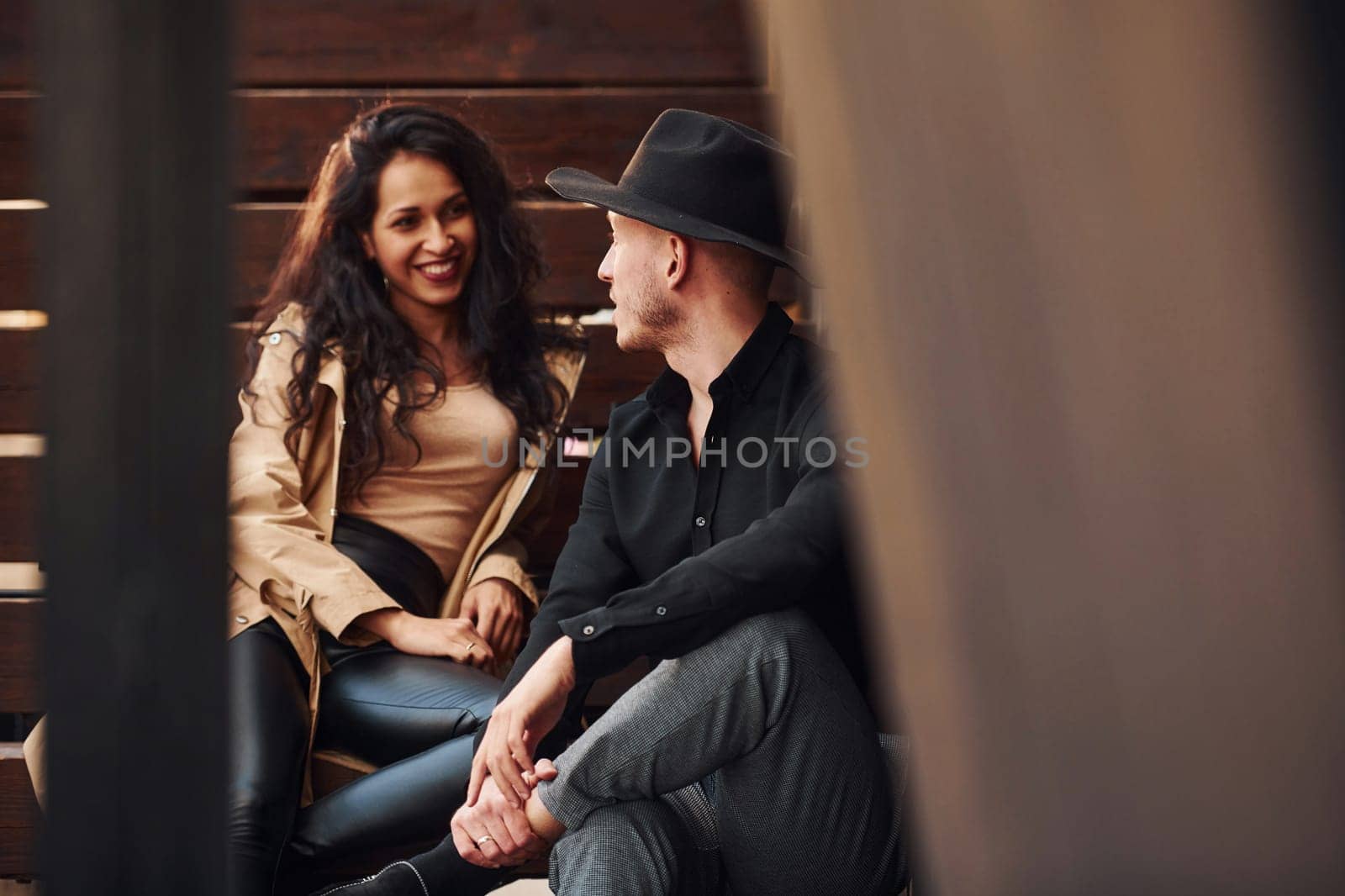 Cheerful brunette in black leggings sitting on wooden stairs with her man in hat.