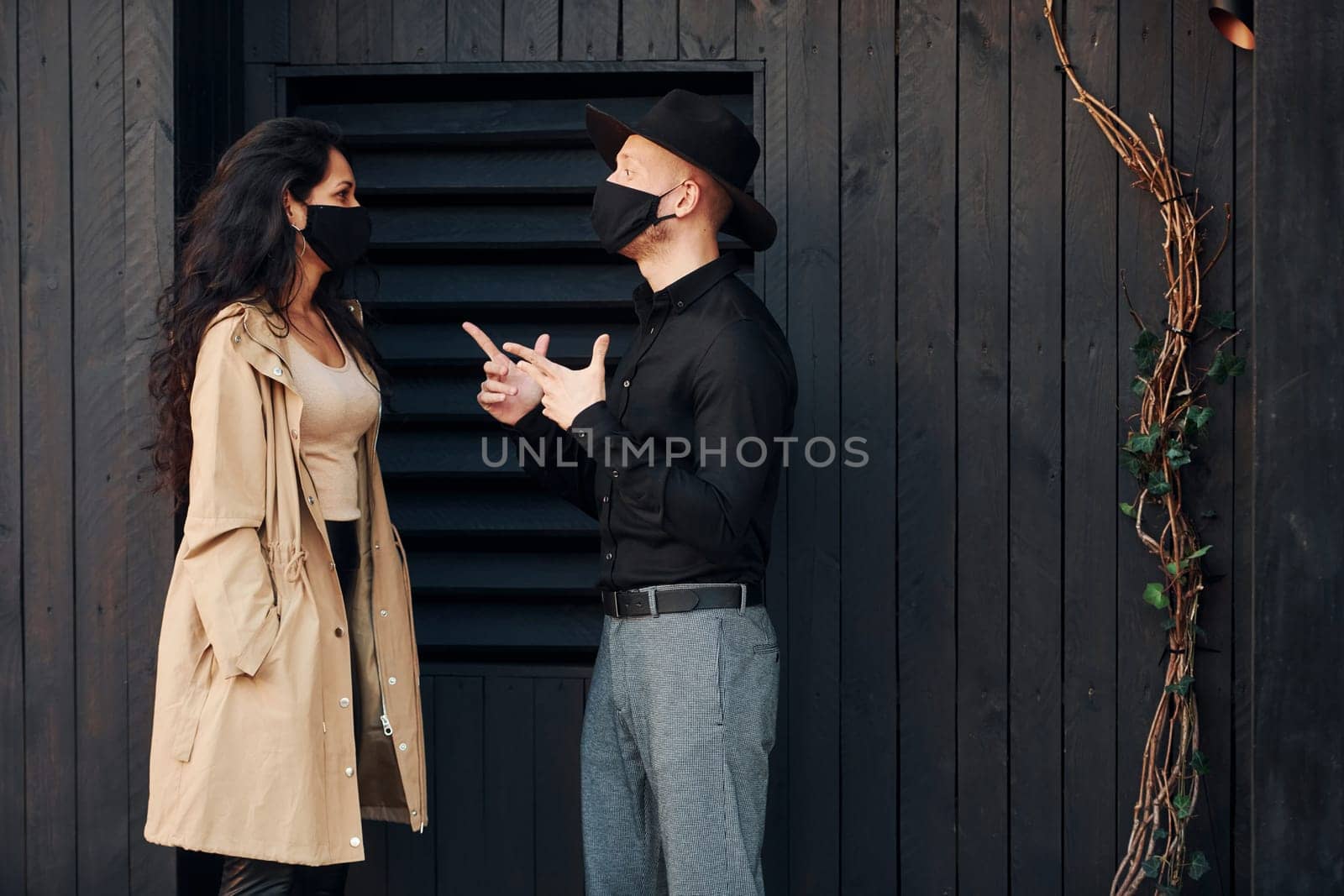 Woman with black curly hair standing against black wooden building exterior with her man in hat by Standret