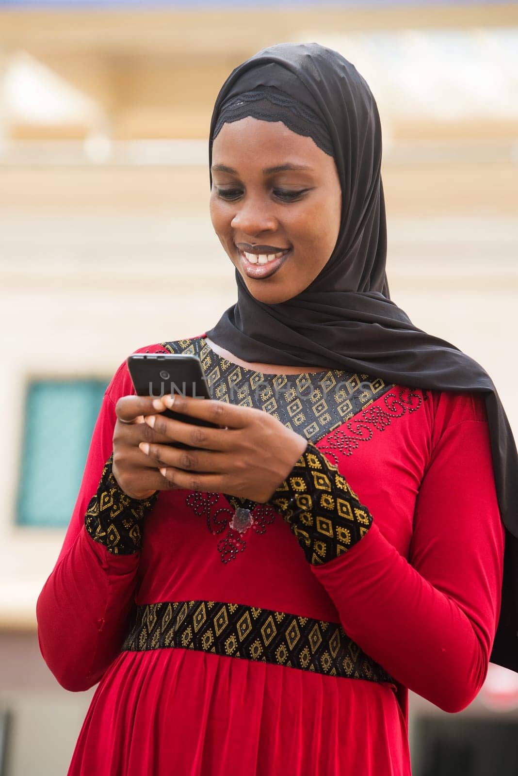 young woman standing outdoors with veiled head looking at mobile phone while smiling.
