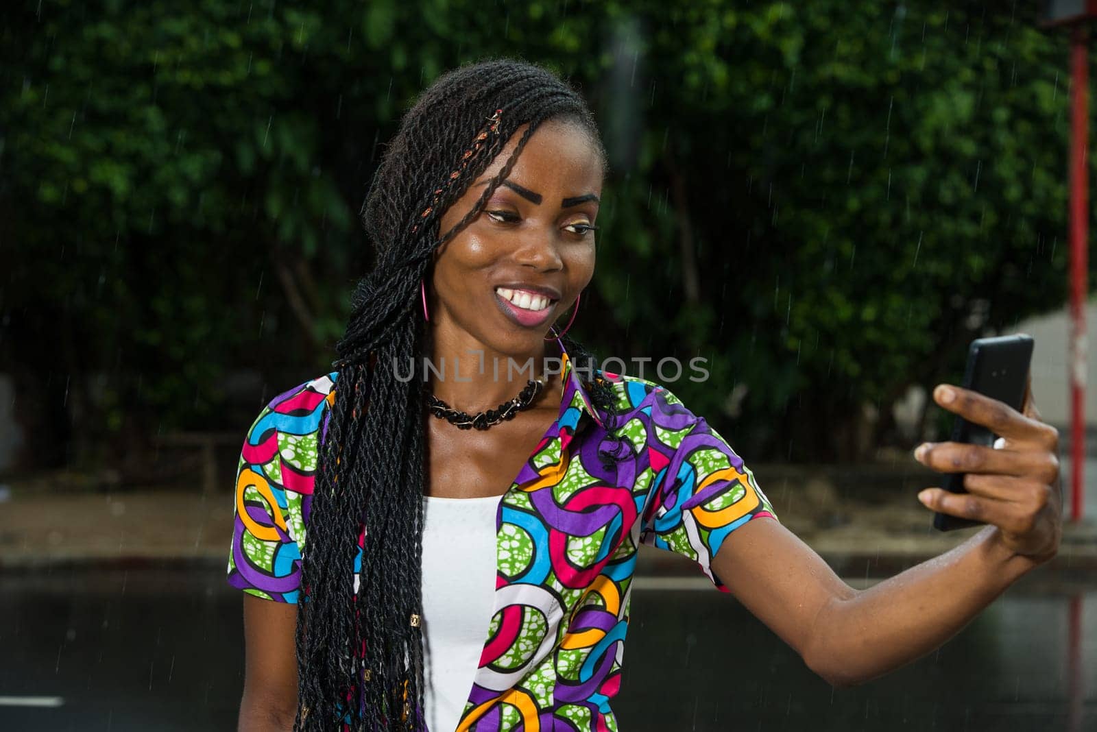 young african girl under a light rain watching mobile phone while laughing.