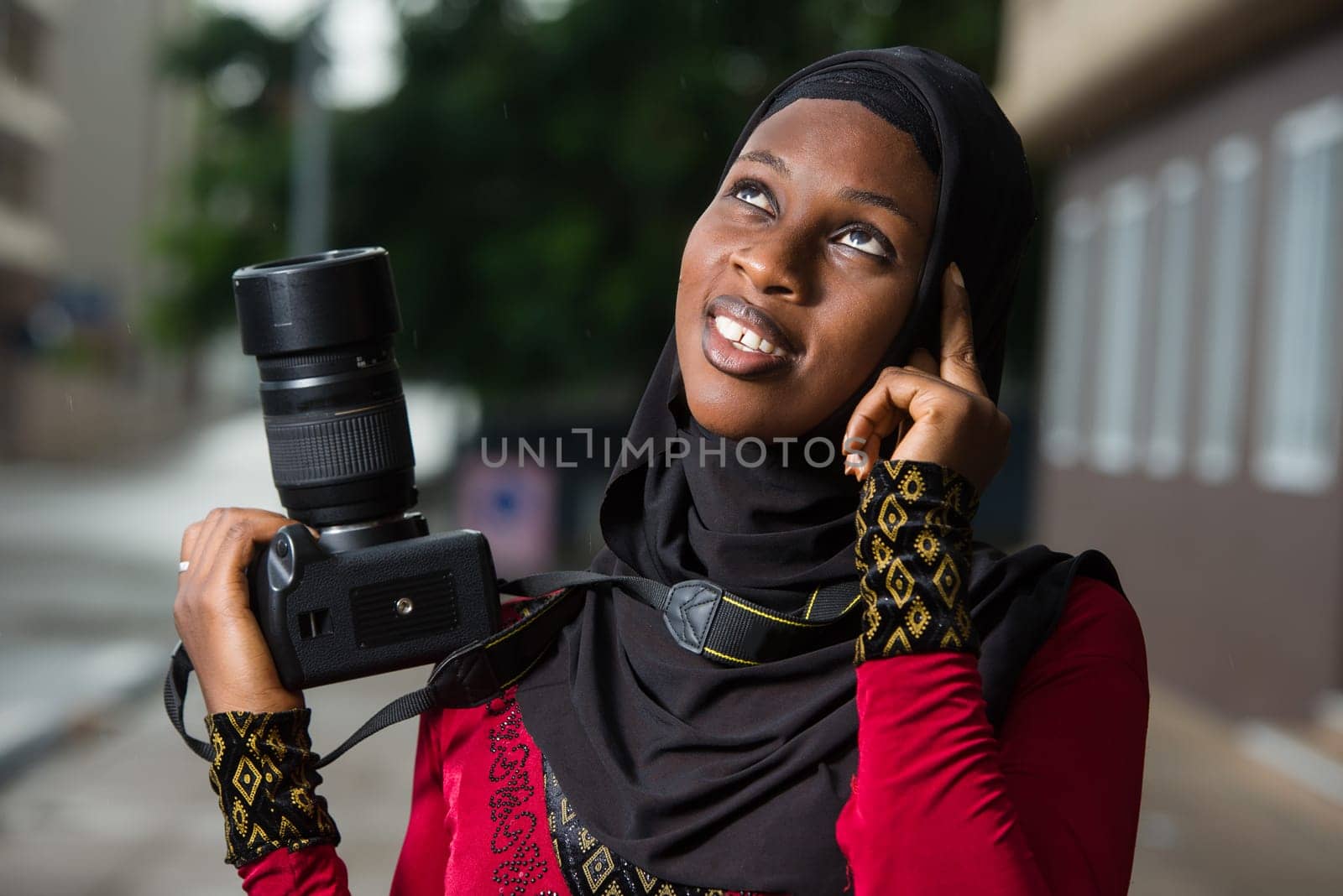 young muslim woman standing outdoors with a camera thinking about looking up there.