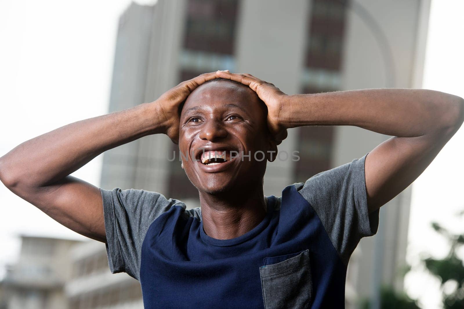 young man standing in t-shirt outdoors laughing with hands on his head.