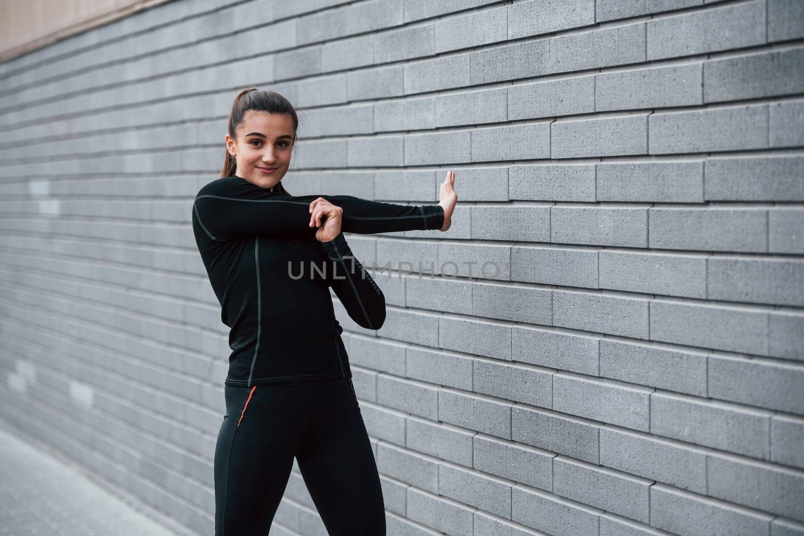 Young sportive girl in black sportswear outdoors doing stretching near gray wall by Standret