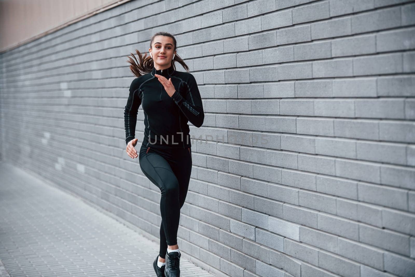 Young sportive girl in black sportswear running outdoors near gray wall by Standret