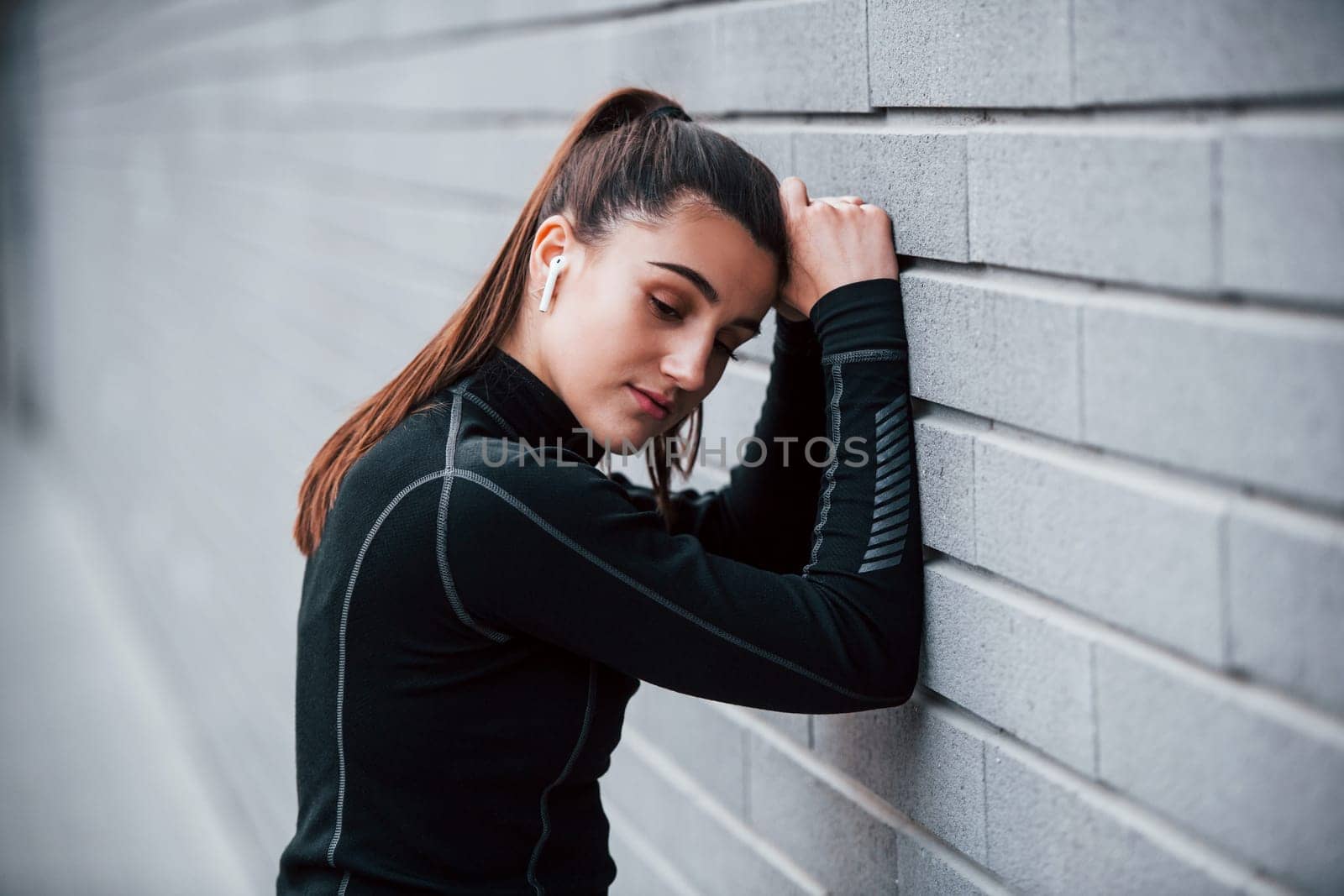 Young sportive girl in black sportswear outdoors near gray wall.