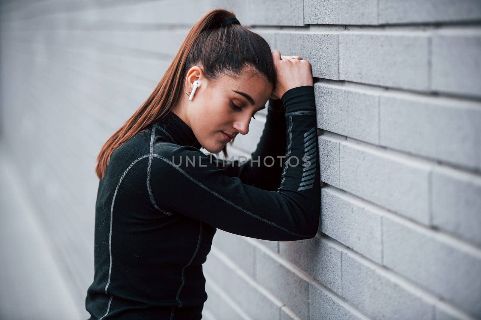 Young sportive girl in black sportswear outdoors near gray wall.