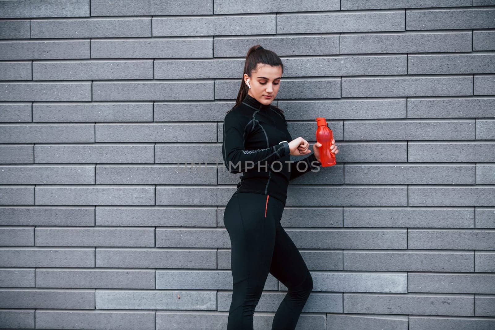 Young sportive girl in black sportswear standing outdoors near gray wall with bottle of water and taking a break.