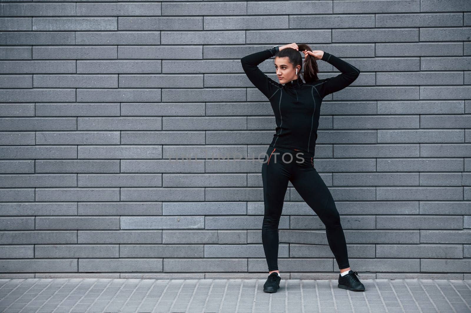 Young sportive girl in black sportswear standing outdoors near gray wall.