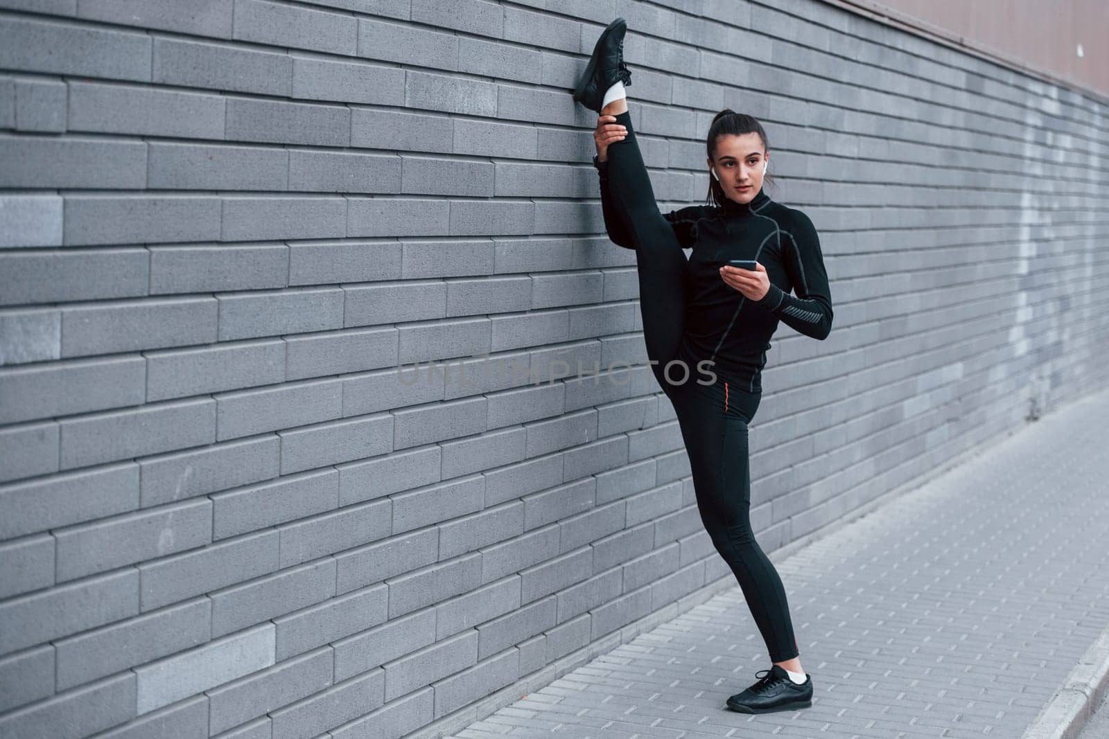 Young sportive girl with phone in black sportswear doing legs stretching outdoors by using gray wall.