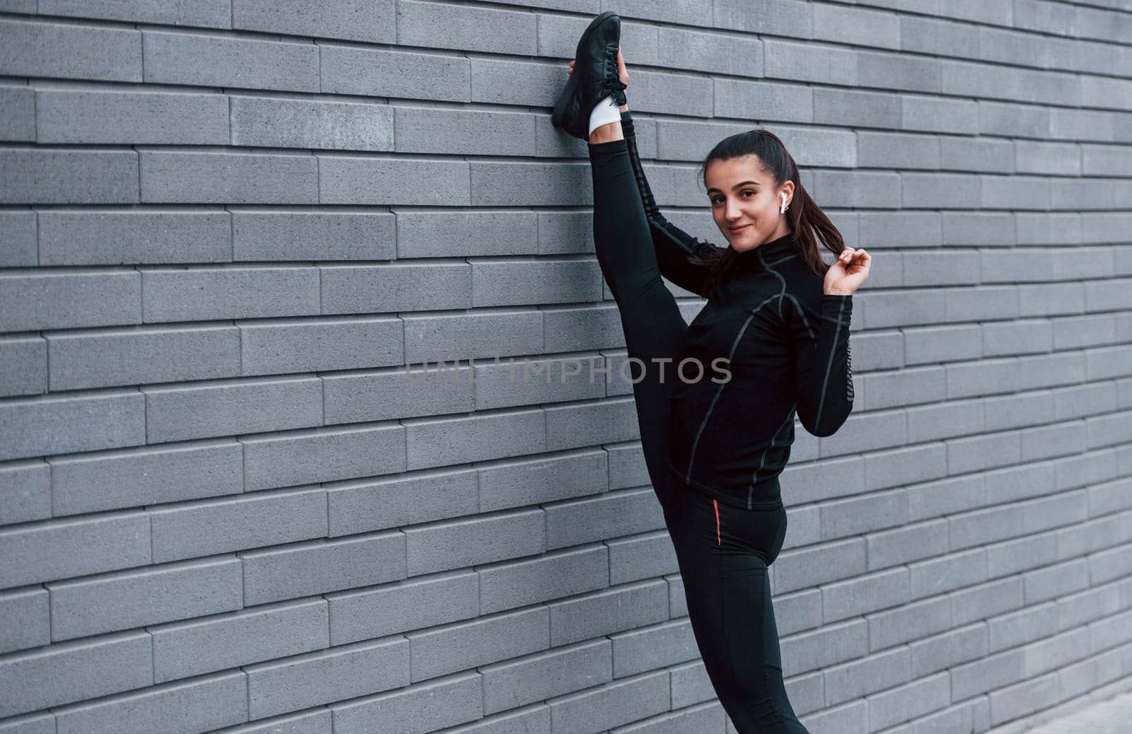 Young sportive girl in black sportswear doing legs stretching outdoors by using gray wall by Standret