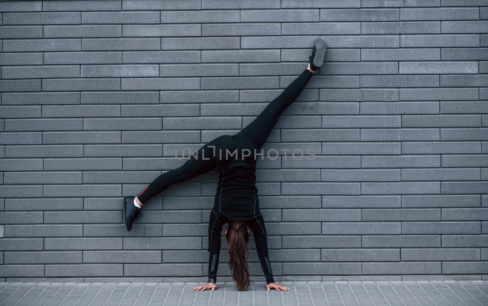 Young sportive girl in black sportswear doing hard handstand exercises outdoors near gray wall by Standret