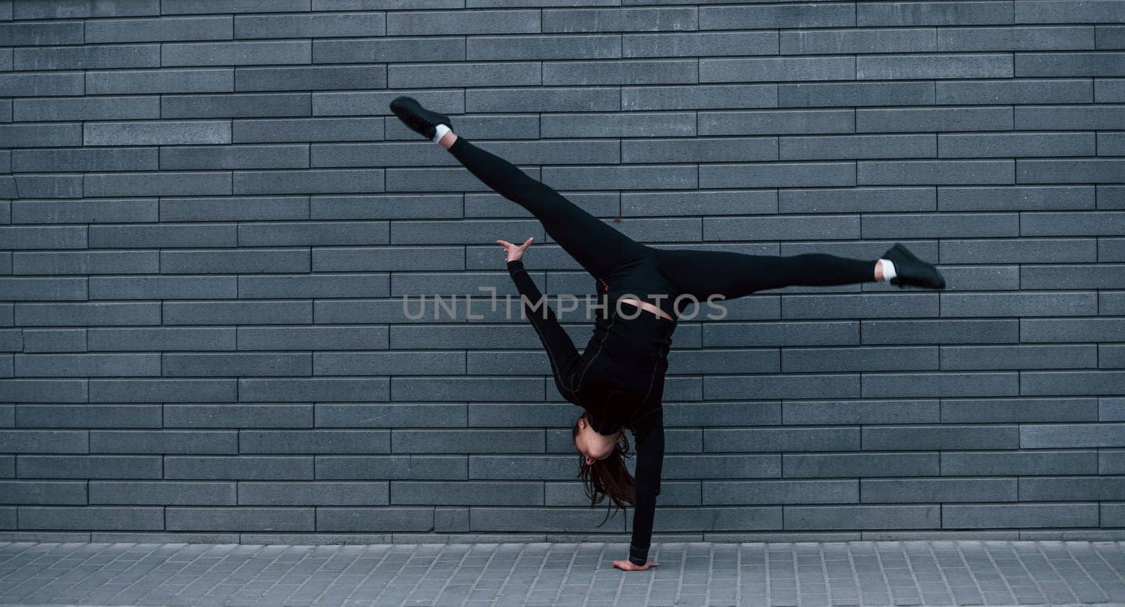 Young sportive girl in black sportswear doing hard handstand exercises outdoors near gray wall.
