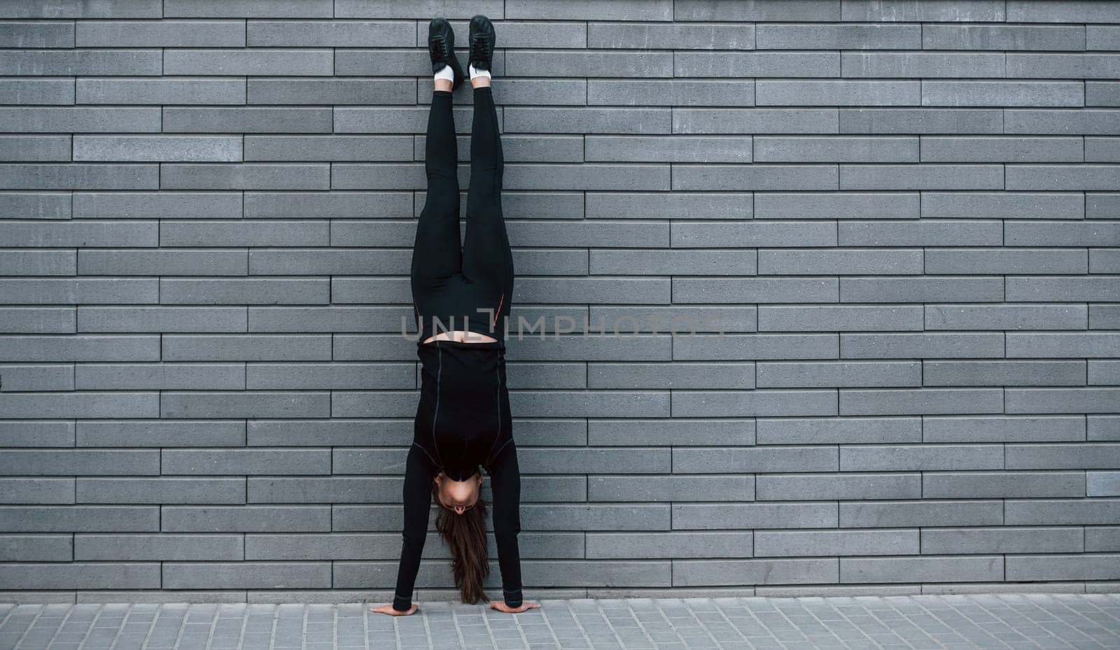 Young sportive girl in black sportswear doing hard handstand exercises outdoors near gray wall.