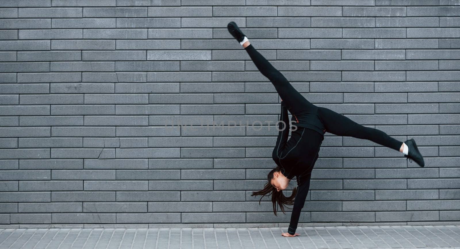 Young sportive girl in black sportswear doing hard handstand exercises outdoors near gray wall by Standret