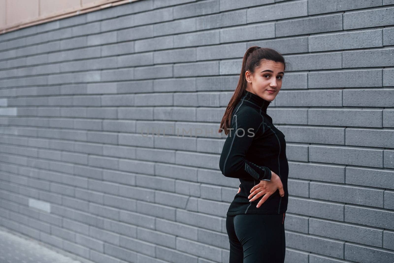 Young sportive girl in black sportswear standing outdoors near gray wall by Standret