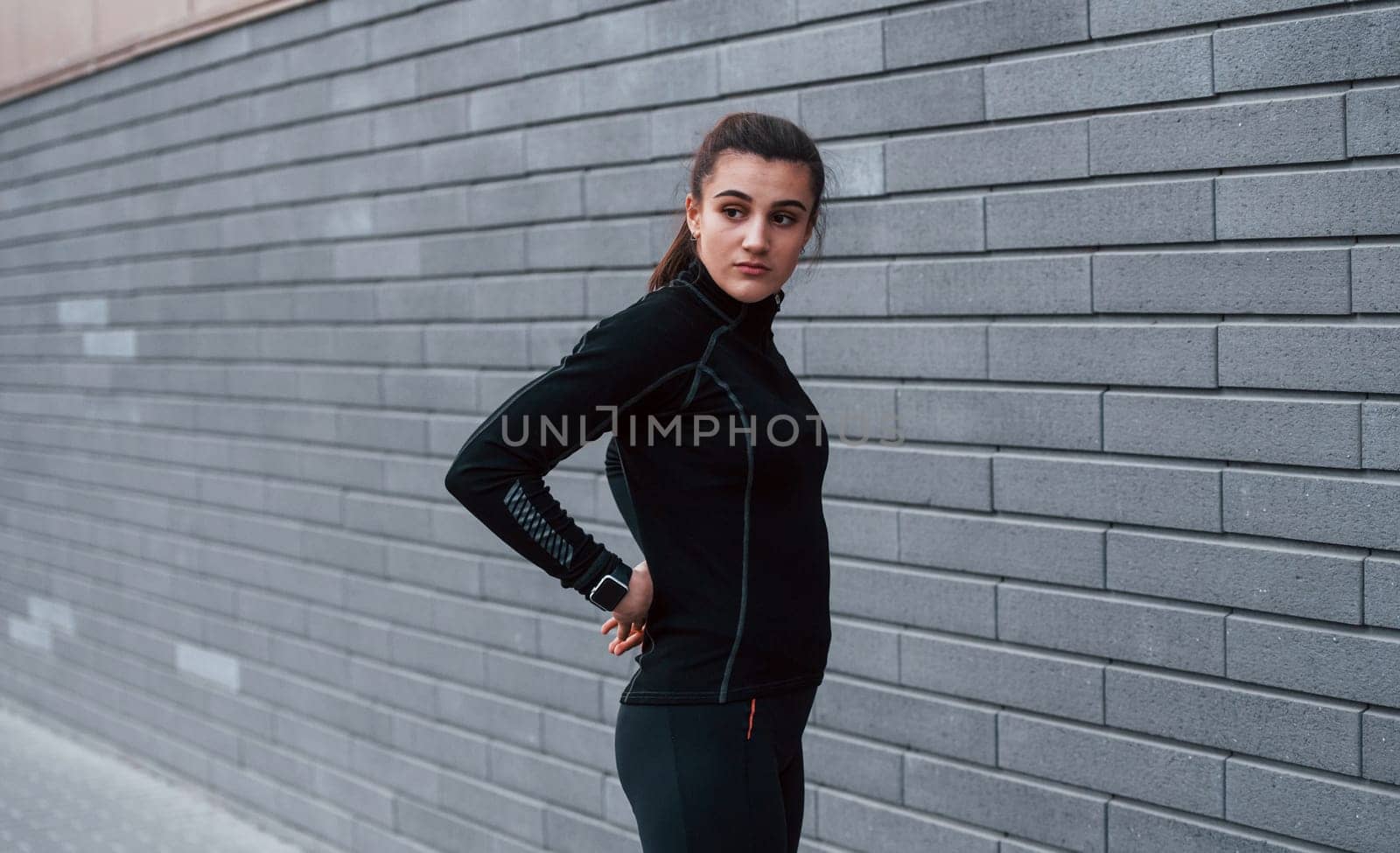 Young sportive girl in black sportswear standing outdoors near gray wall by Standret