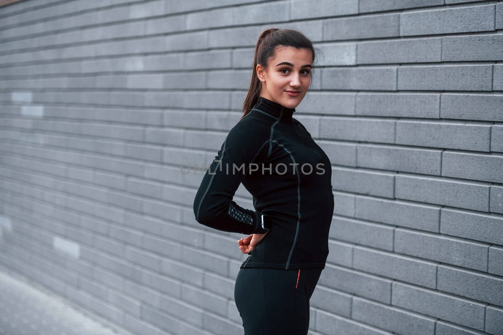 Young sportive girl in black sportswear standing outdoors near gray wall.