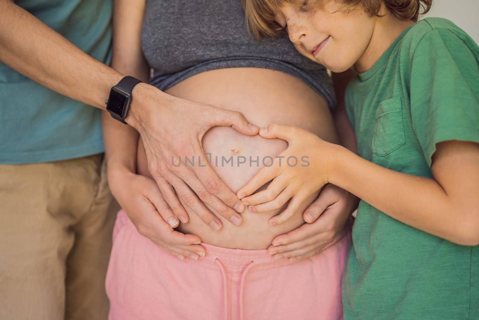 Pregnant Woman, father and first kid child holding hands in a heart shape on her baby bump. Pregnant Belly with fingers Heart symbol.
