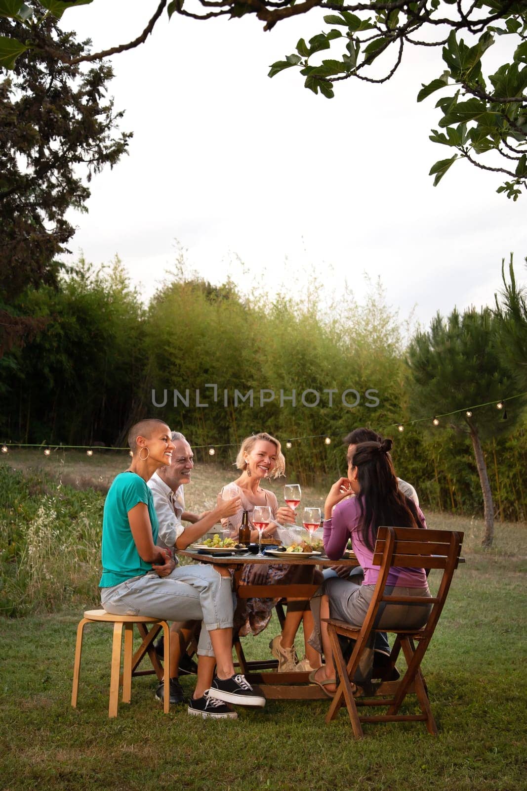 Happy friends laughing and eating at sunset during garden dinner party. Vertical image. Copy space. Lifestyle.