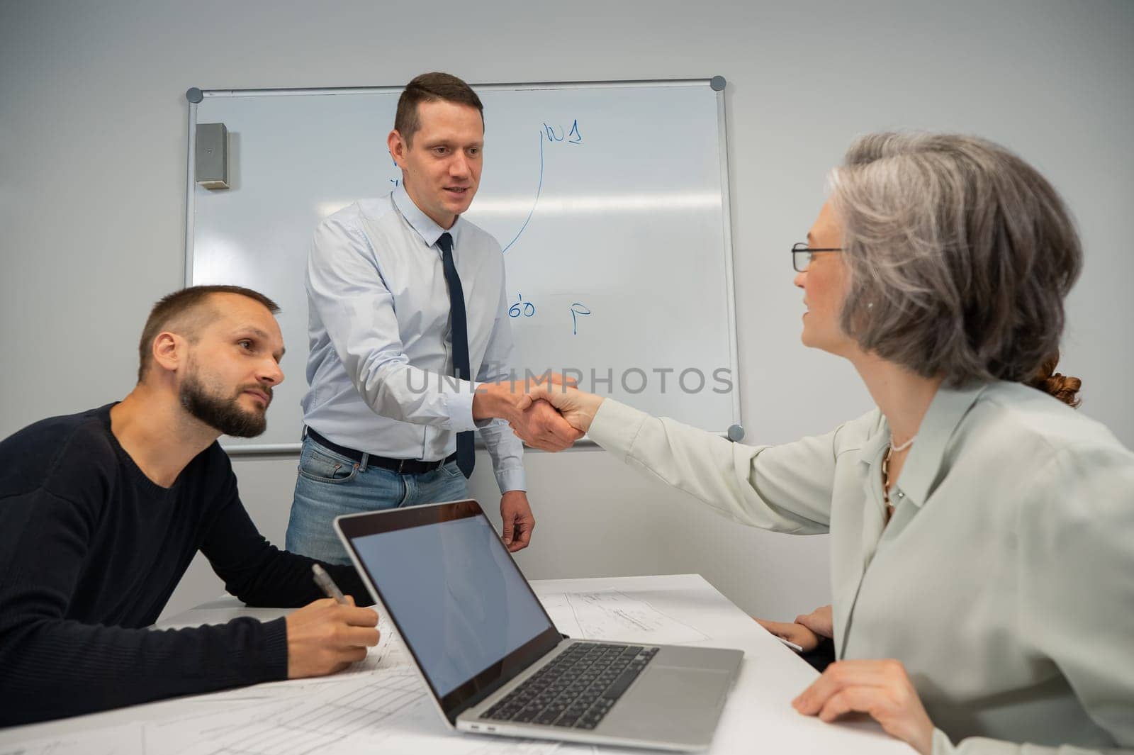 The boss makes a presentation to subordinates at the white board. Caucasian man shaking hands with middle aged woman. by mrwed54