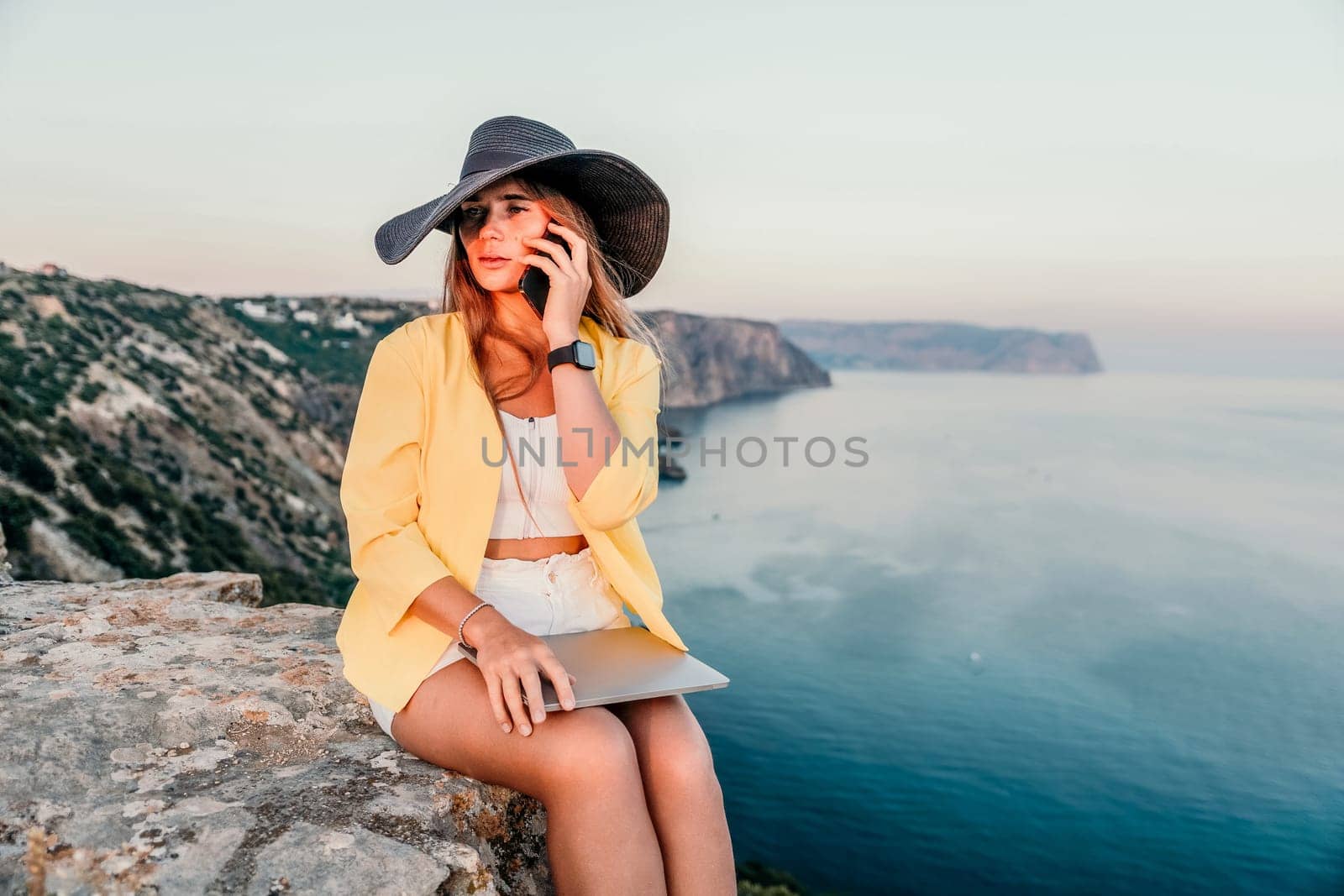 Successful business woman in yellow hat working on laptop by the sea. Pretty lady typing on computer at summer day outdoors. Freelance, travel and holidays concept.