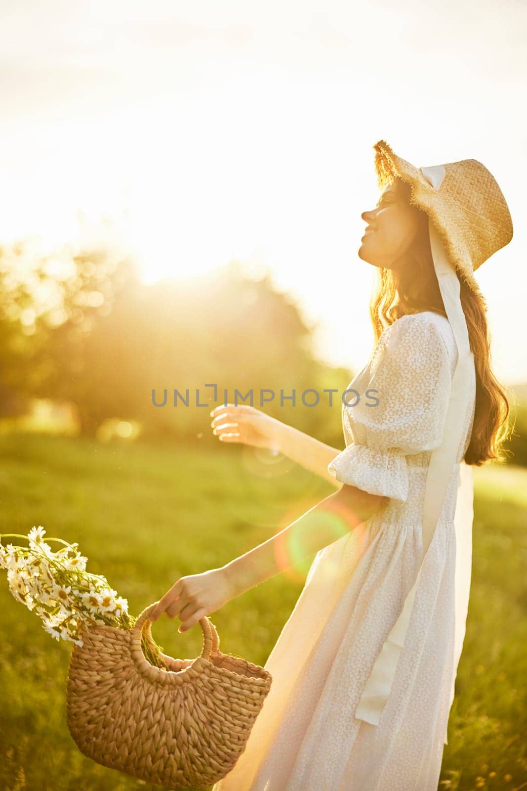 a beautiful woman in a hat and a light dress walks through a chamomile field with a basket full of daisies in the rays of the setting sun. High quality photo