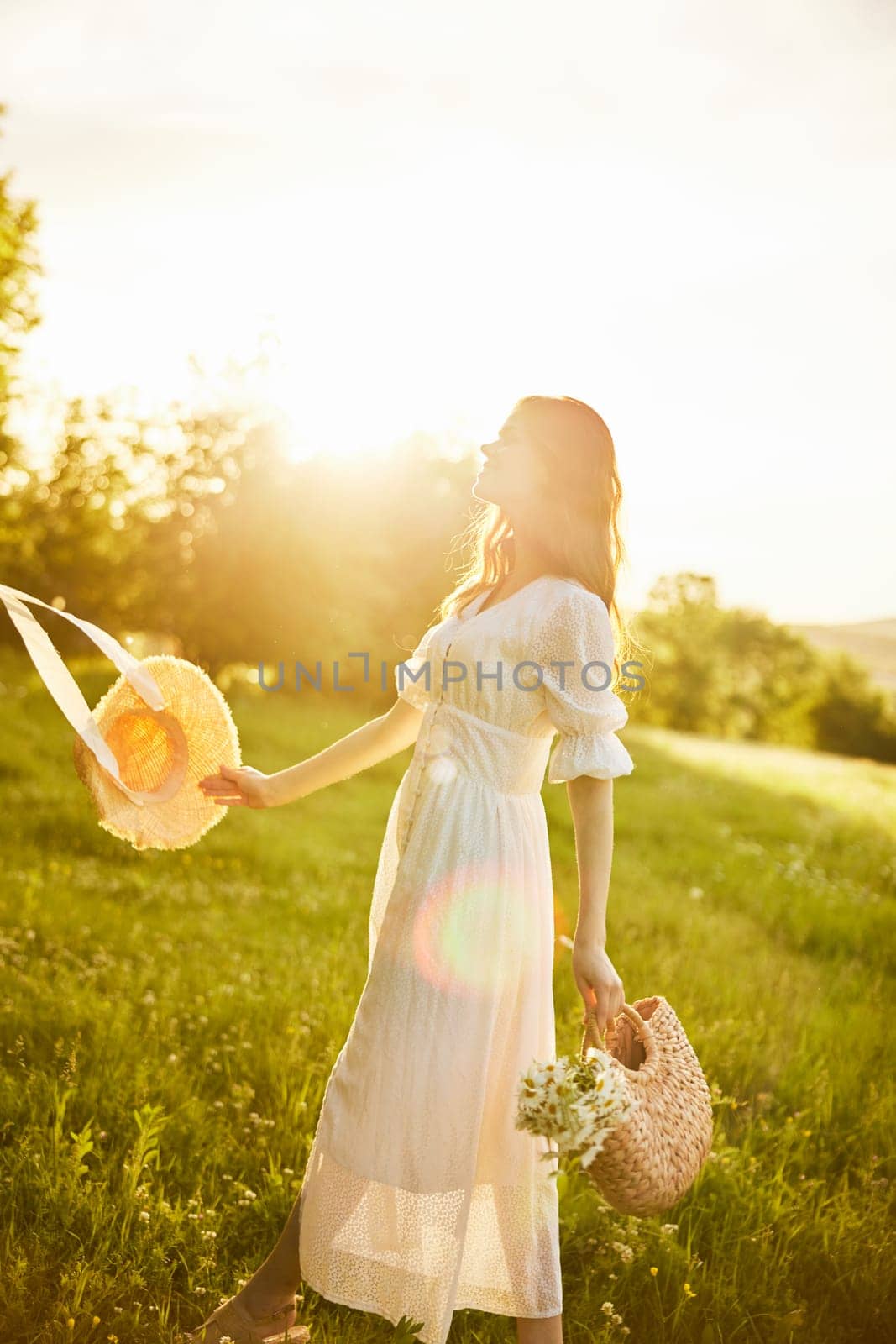 a beautiful woman in a hat and a light dress walks through a chamomile field with a basket full of daisies in the rays of the setting sun by Vichizh