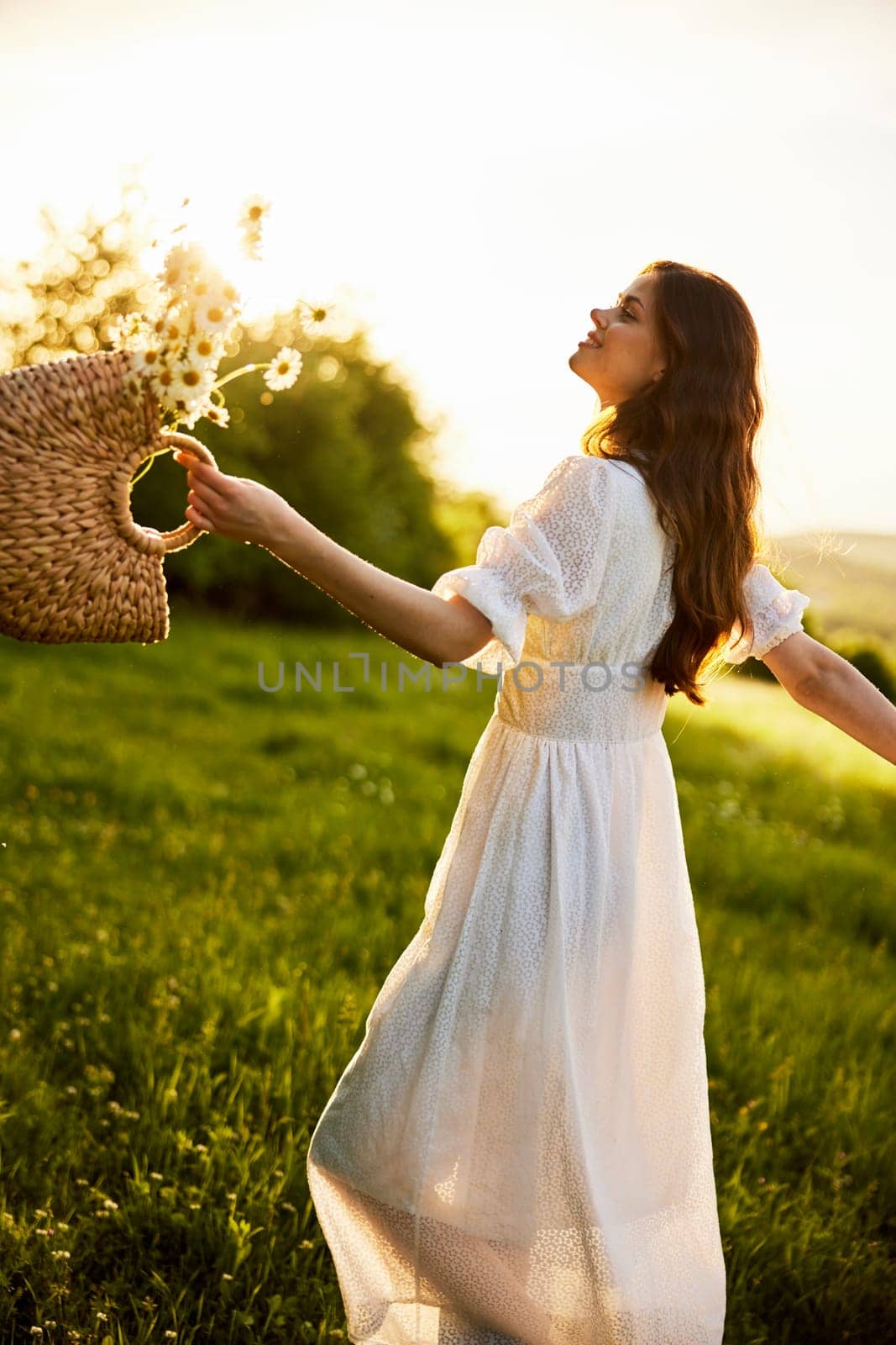 a beautiful woman in a hat and a light dress walks through a chamomile field with a basket full of daisies in the rays of the setting sun by Vichizh