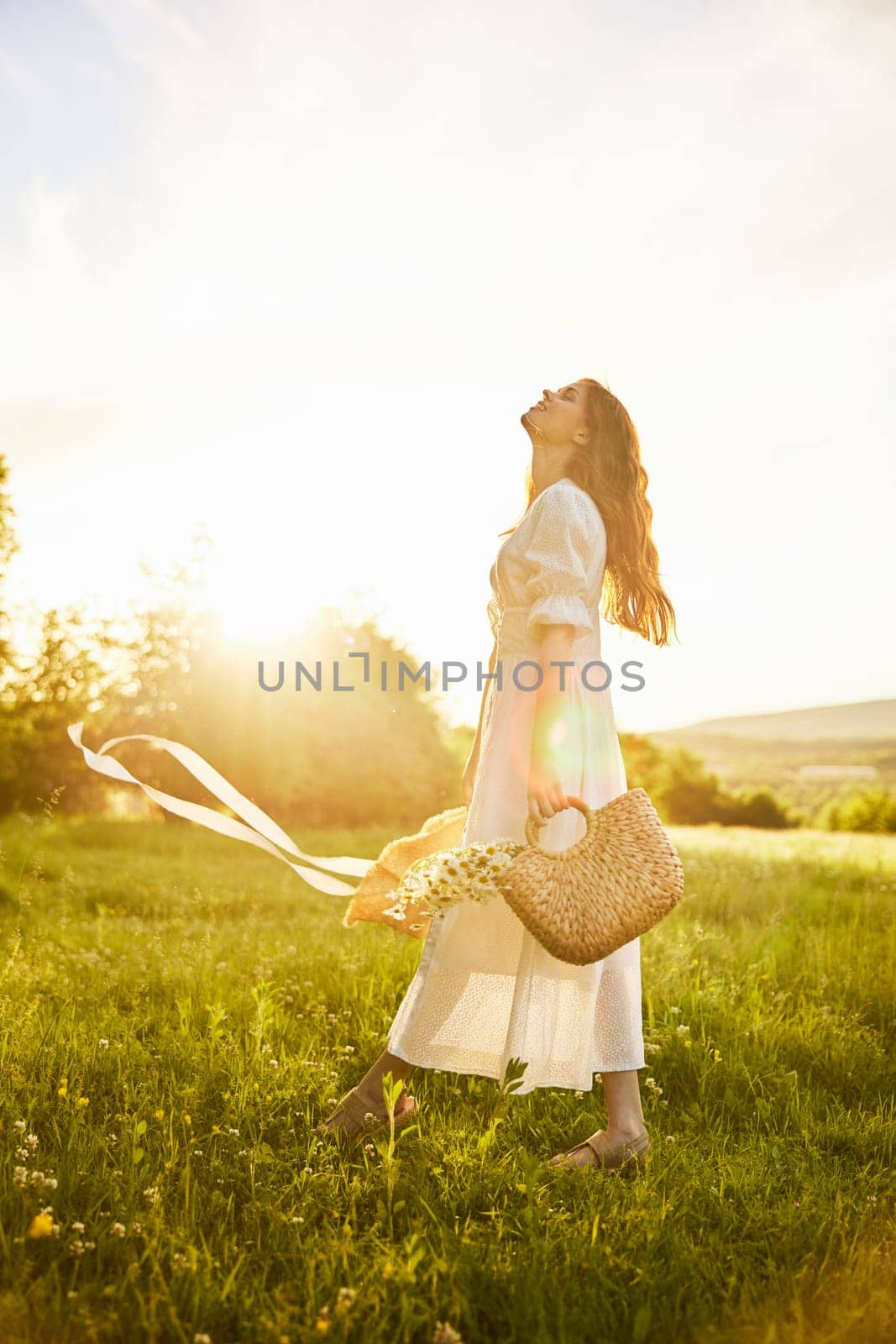 a beautiful woman in a hat and a light dress walks through a chamomile field with a basket full of daisies in the rays of the setting sun. High quality photo