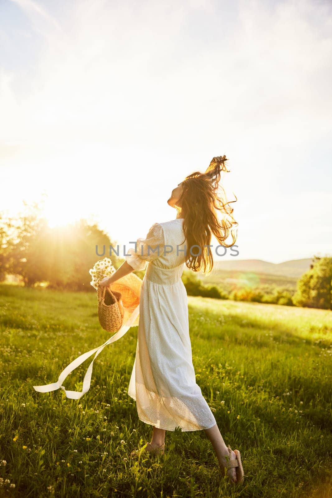 a woman in a light dress in a chamomile field against the background of a sunset stands with her back to the camera with a basket of flowers in her hands by Vichizh