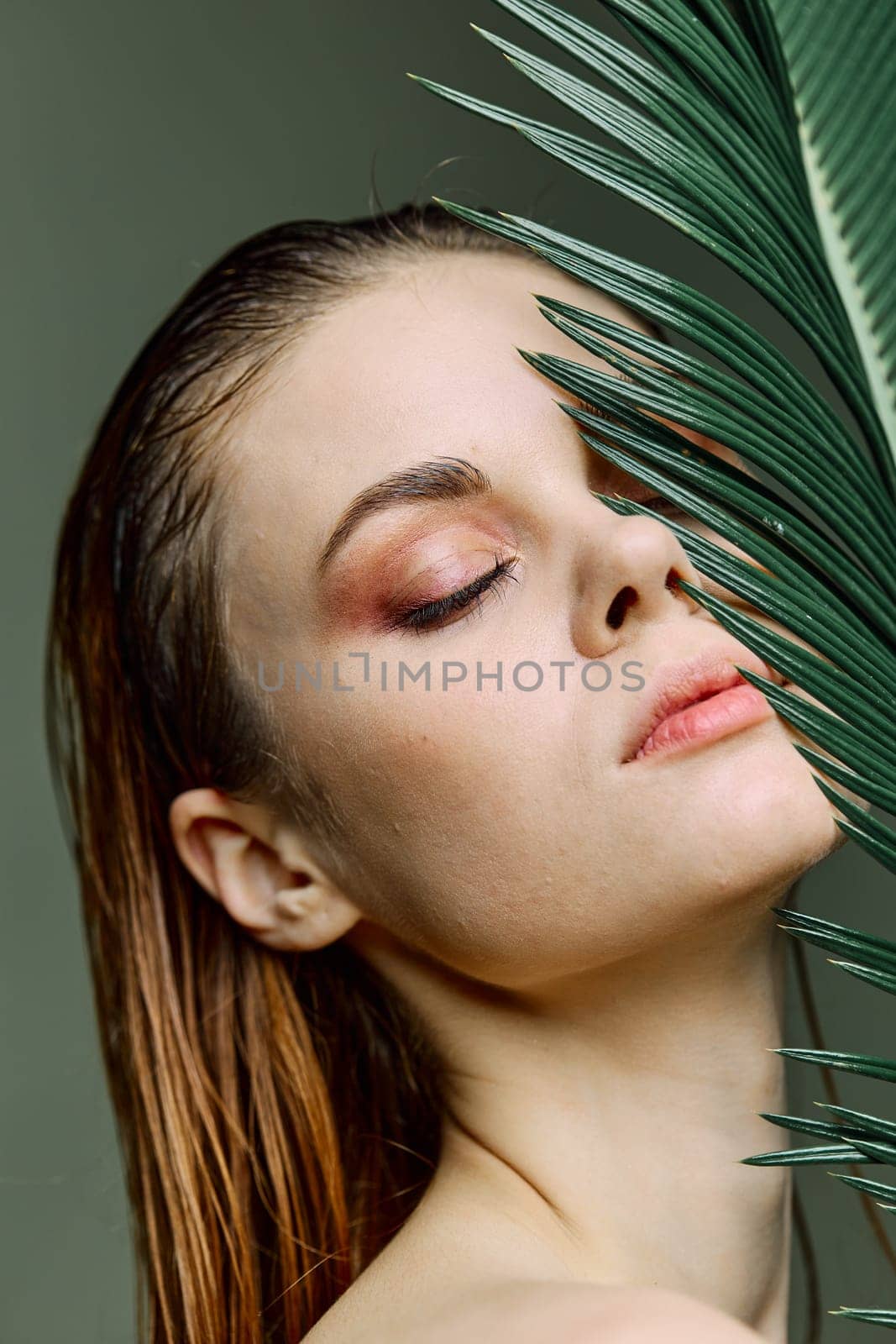 a close beauty portrait of a beautiful woman standing holding a palm leaf near her face, closing her eyes. Vertical photo without retouching of problem skin. High quality photo