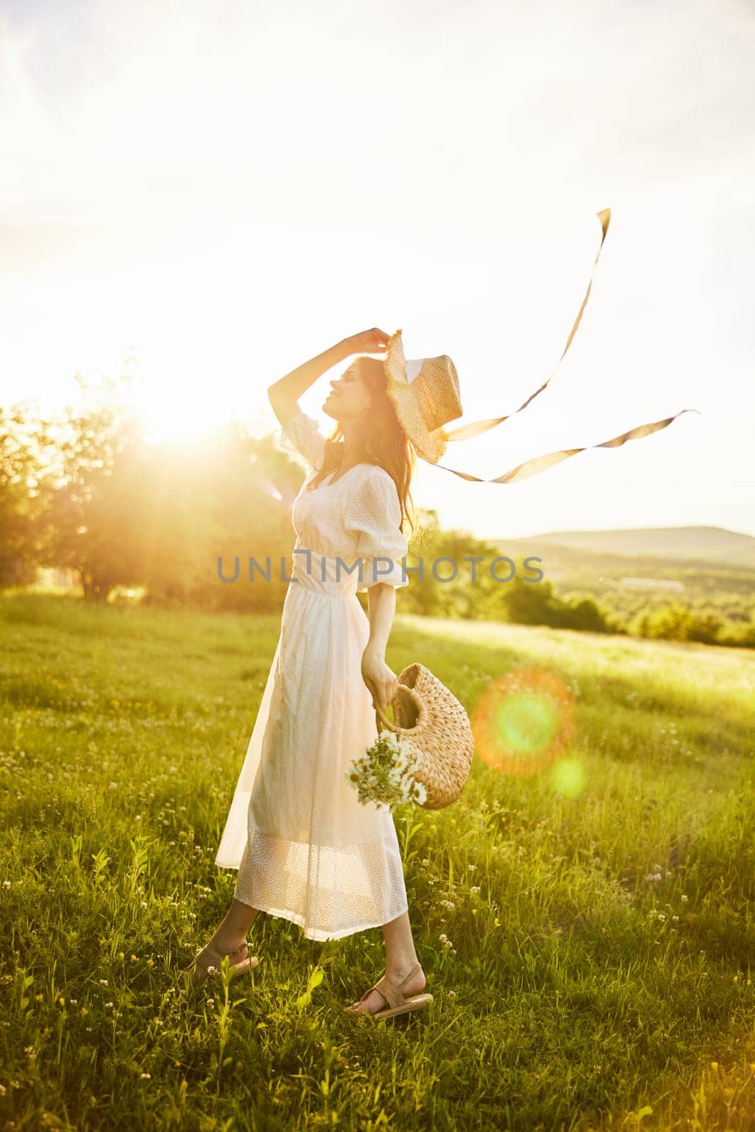 a beautiful woman in a hat and a light dress walks through a chamomile field with a basket full of daisies in the rays of the setting sun by Vichizh
