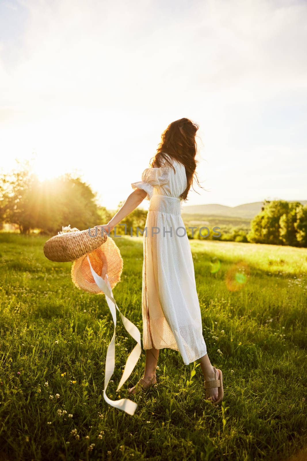 a woman in a light dress in a chamomile field against the background of a sunset stands with her back to the camera with a basket of flowers in her hands by Vichizh
